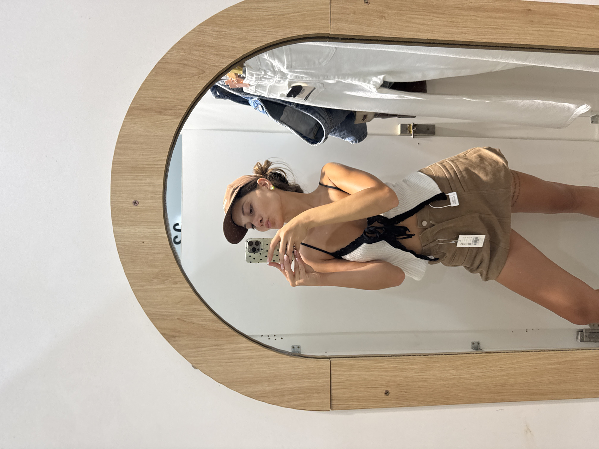 A woman in a changing room takes a selfie in front of a wooden-framed mirror. She wears a brown baseball cap, a white tank top with black straps, and brown shorts with tags. Above her are hung articles of clothing, including white shirts and a blue denim jacket. The changing room's walls are white. The image was taken in a fitting room; there is no clear evidence of Đà Nẵng, Vietnam.