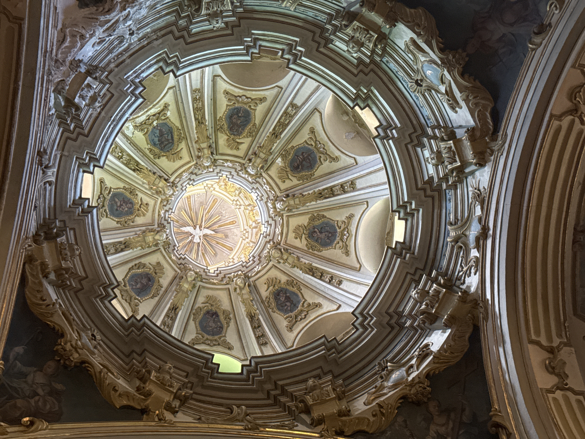 The image is a low-angle, interior shot looking up at the dome of the Sant Bartomeu church in Sóller, Spain. The dome is ornate, featuring painted panels depicting cherubs within gilded frames and a central opening with radiating golden beams of light. The architecture of the dome's supports and decorative elements are highly detailed, with carved gold and white trim. The overall impression is of opulent religious architecture.