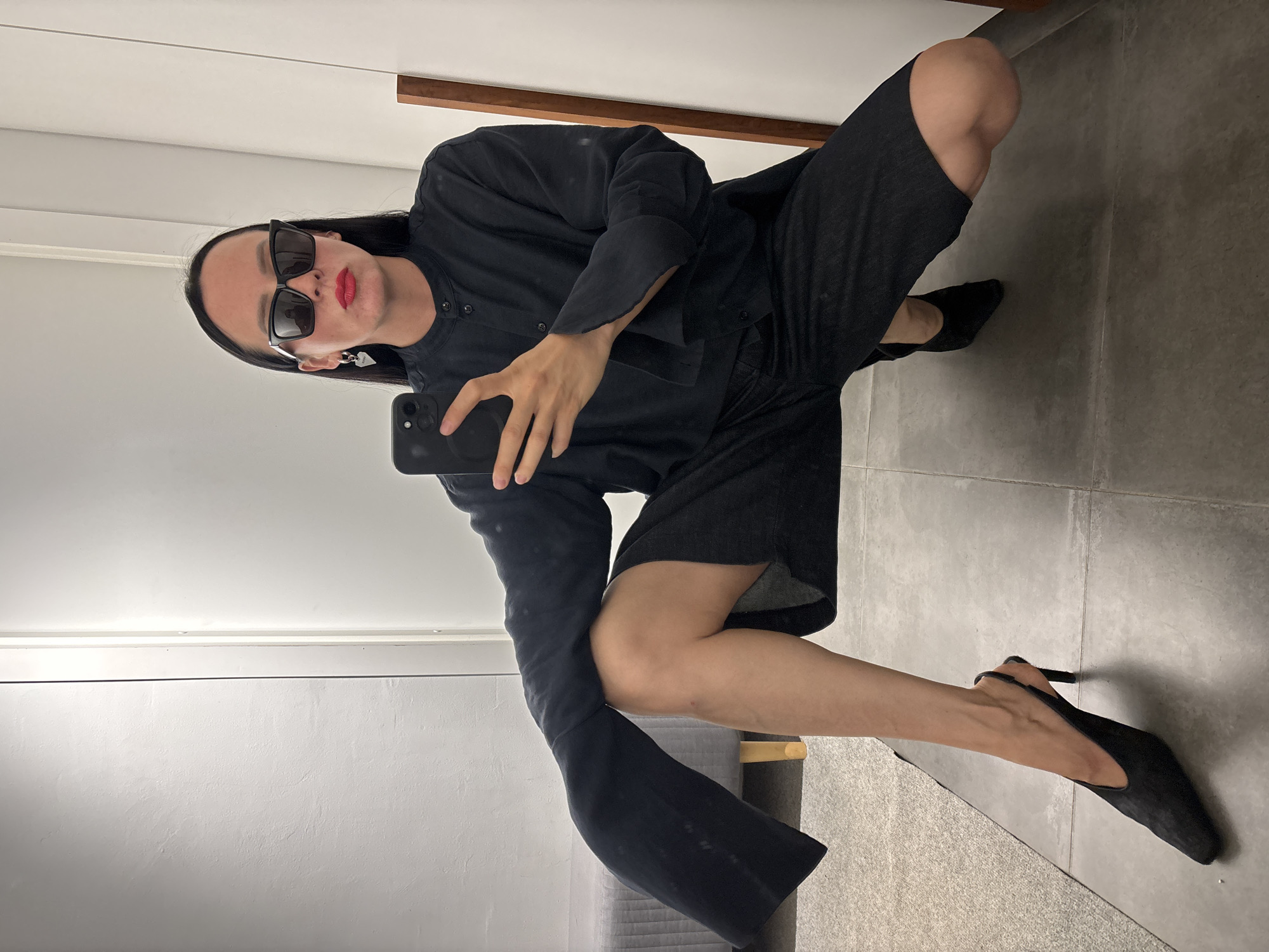 A person wearing sunglasses, black clothing, and black heels is crouching in what appears to be a room with gray tile flooring and white walls. The person is holding a smartphone, seemingly taking a selfie. A section of wooden trim and a gray upholstered object are visible in the background. The exact location in Chaikovsky, Russia cannot be determined from the image.