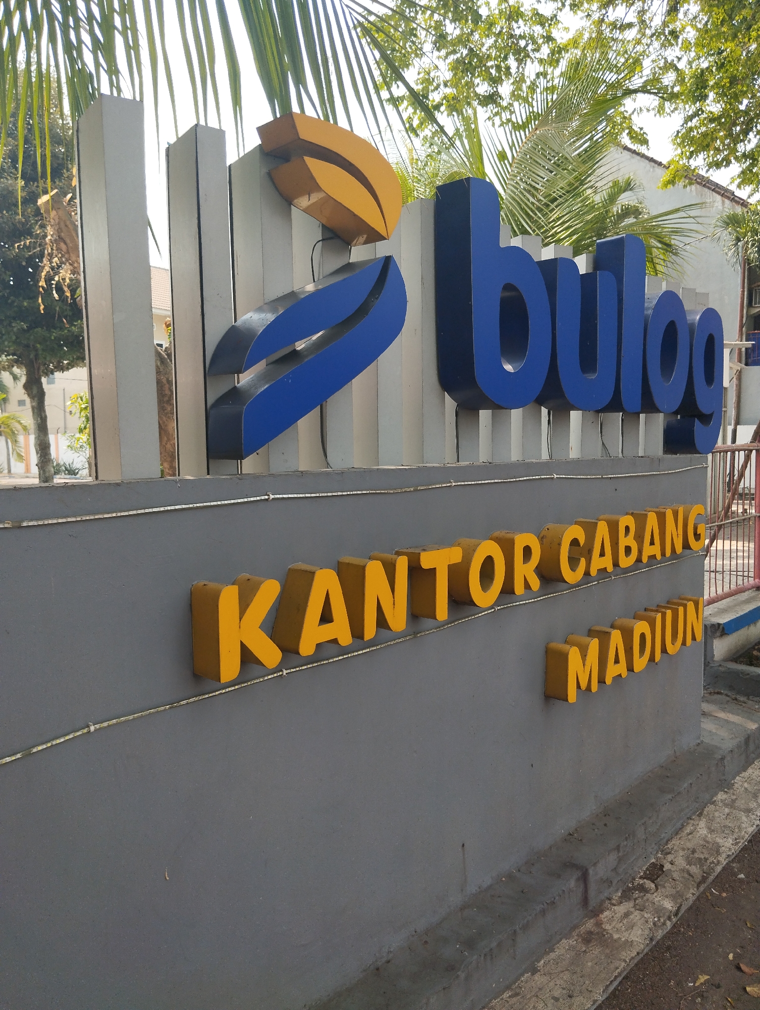 The image displays the exterior signage of a Bulog branch in Madiun, Indonesia. The prominent sign features the "Bulog" logo, which consists of blue curved elements and a golden curved shape above it. Below the logo, a gray concrete wall displays three-dimensional yellow lettering spelling out "KANTOR CABANG MADIUN," meaning "Madiun Branch Office." The sign is situated outdoors, with lush green foliage and trees visible in the background, suggesting a sunny day. The foreground shows a concrete pathway and some fallen leaves. The overall scene is that of a government or public institution's entrance.