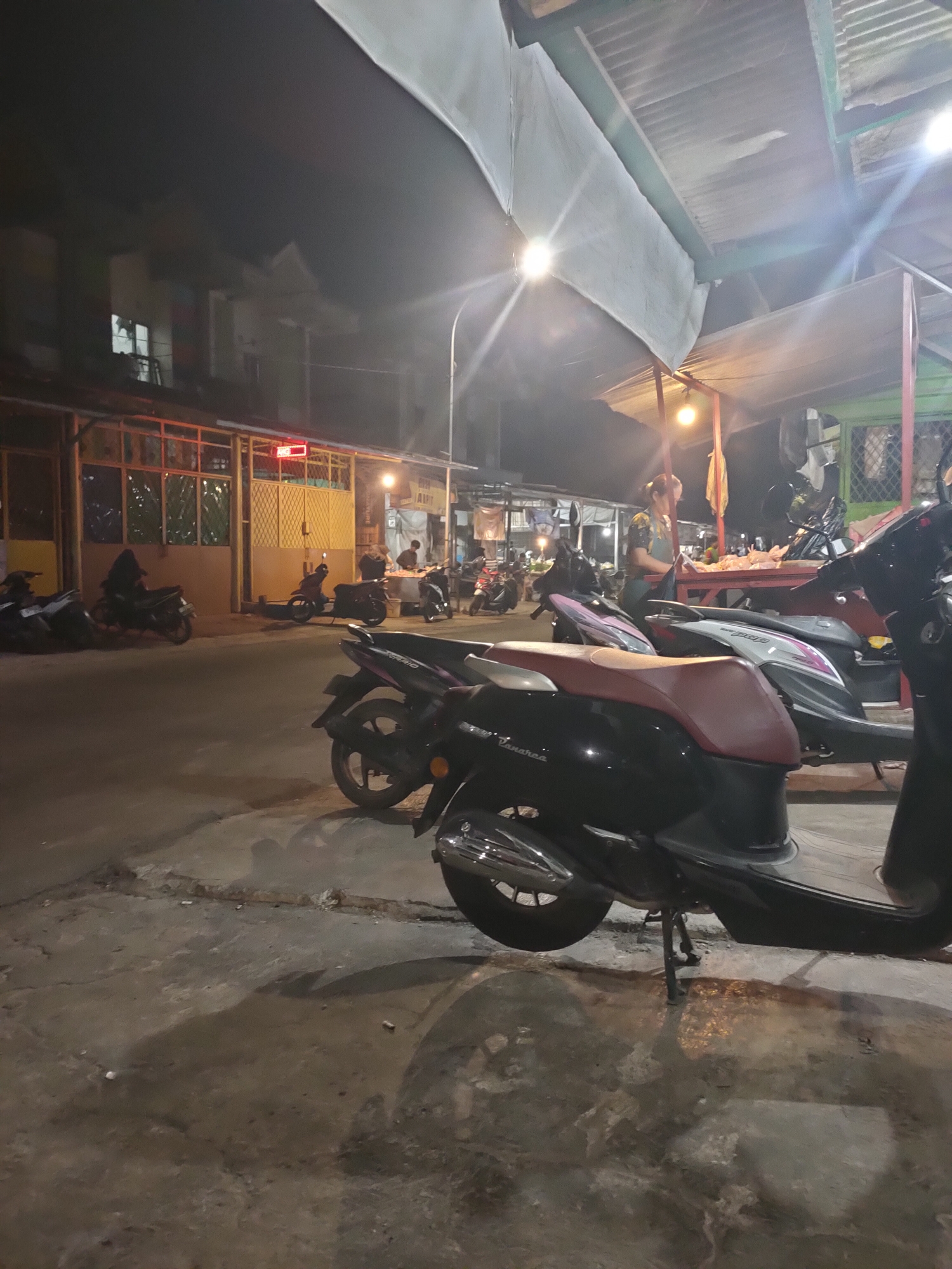 A nighttime, outdoor street scene in Subang, Indonesia, features a row of parked motorcycles in the foreground, including a black and red scooter prominently displayed. Behind the scooters, a woman in a dark top and apron stands at a red market stall, appearing to be tending to goods. Several other motorcycles are parked further down the street, and people are visible near the stalls. The background shows buildings with illuminated windows and a covered structure with corrugated metal roofing, illuminated by overhead lights. The ground is paved and shows some signs of wear.