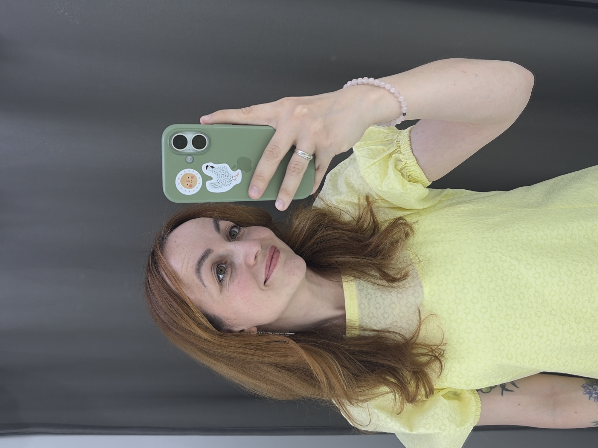 A woman is taking a selfie with a green phone. She has auburn hair and is wearing a yellow dress with puffed sleeves. She also has a pink beaded bracelet. The background appears to be a gray wall and a white surface. The location is Chaikovsky, Russia.