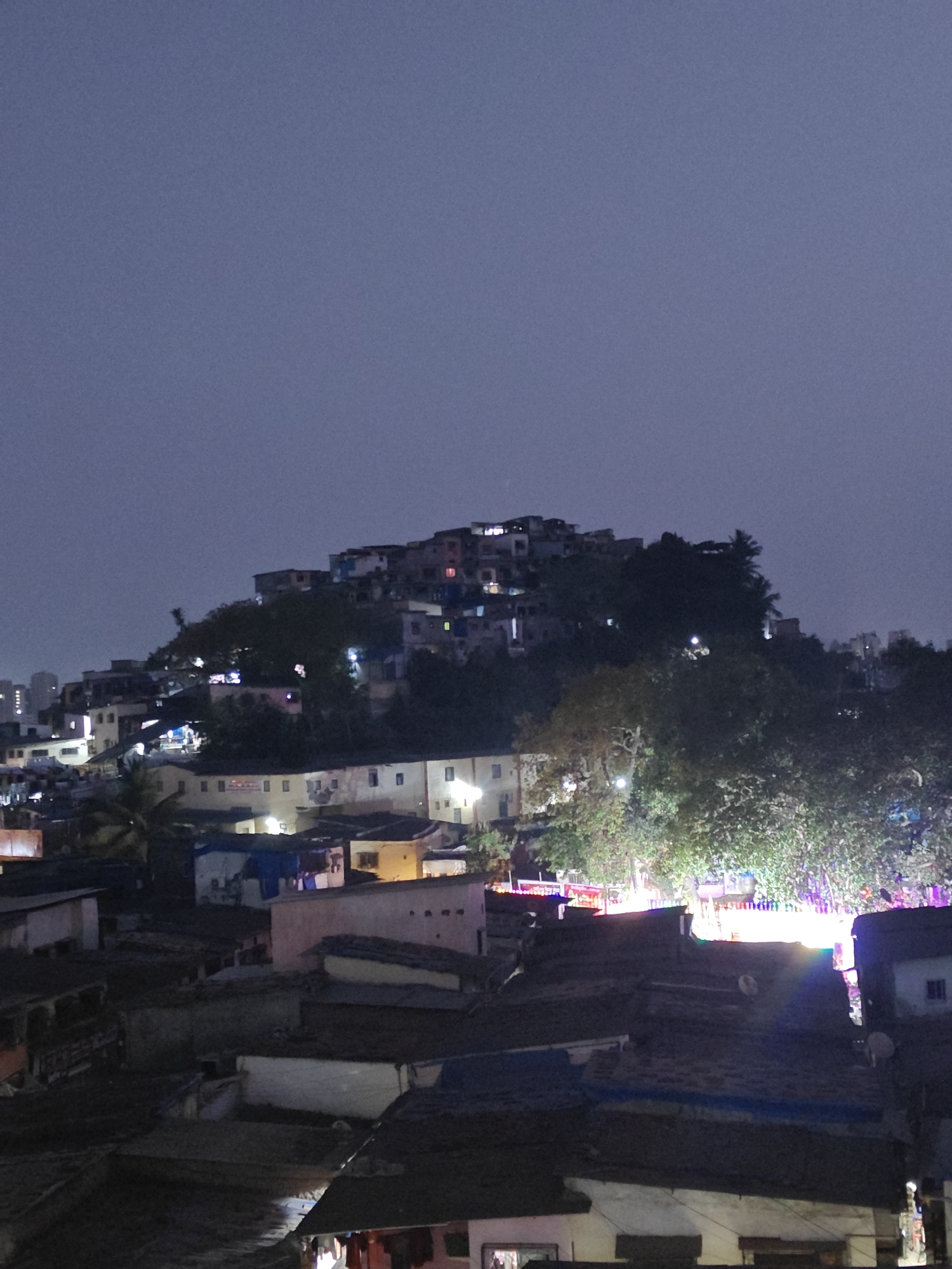 The image depicts a nighttime scene in Mumbai, India, showing a densely populated hillside slum area with a mix of buildings, vegetation, and lights. The structures appear to be tightly packed together, ranging from smaller shacks to slightly larger, more established buildings. Lights illuminate the buildings and streets, indicating human activity. The area is situated on a hill, with buildings cascading down the slope. The sky is dark, suggesting it is late evening or night. In the foreground, rooftops of the lower dwellings are visible. The overall impression is one of urban density and activity.