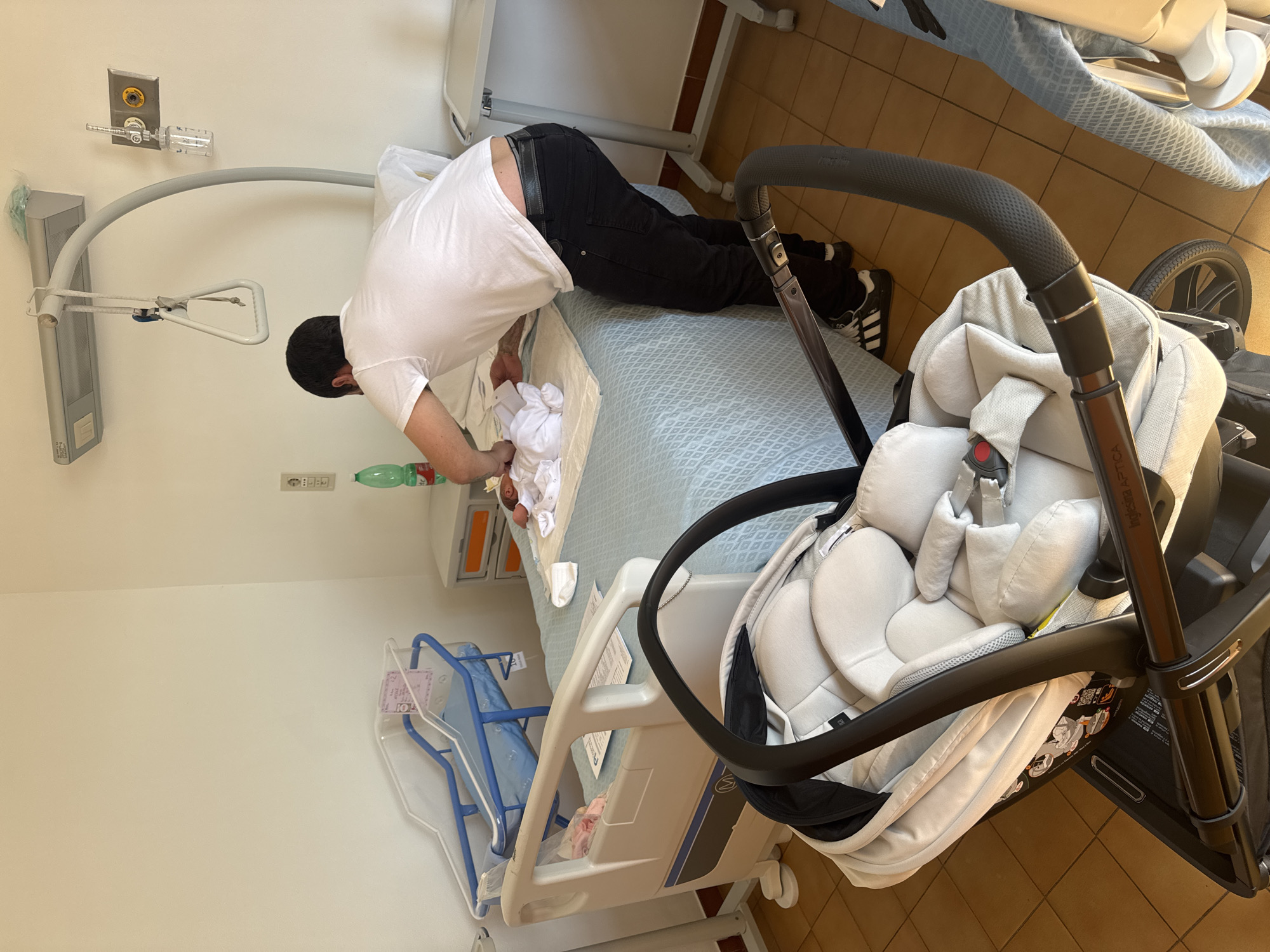 This image captures a tender moment in what appears to be a hospital or birthing center room in Aprilia, Italy. The central subject is a man, dressed in a white t-shirt, black jeans, a black belt, and black and white sneakers, leaning over a baby lying on a hospital bed. The baby is small and wrapped in white fabric, being attended to by the man, possibly during a changing or comforting moment.
The setting is bright, suggesting daytime. The room features a hospital bed with a blue patterned sheet, and a clear plastic baby bassinet on wheels is visible to the left. On the wall, medical equipment including oxygen or gas outlets and a flow meter are mounted. A bed mobility aid, commonly known as a trapeze bar, is attached to the bed frame. A green plastic water bottle is on the floor next to the bed.
In the foreground and to the right, a modern baby carriage or stroller is prominent. It features a black frame and light grey, padded fabric, characteristic of an infant car seat attached to a stroller base. Text visible on the stroller reads "Inglesina Aptica." Another faint number, possibly "101," can be seen on an item within the baby bassinet. The floor is tiled in a light brown pattern. The overall scene conveys a sense of new parenthood in a medical environment.