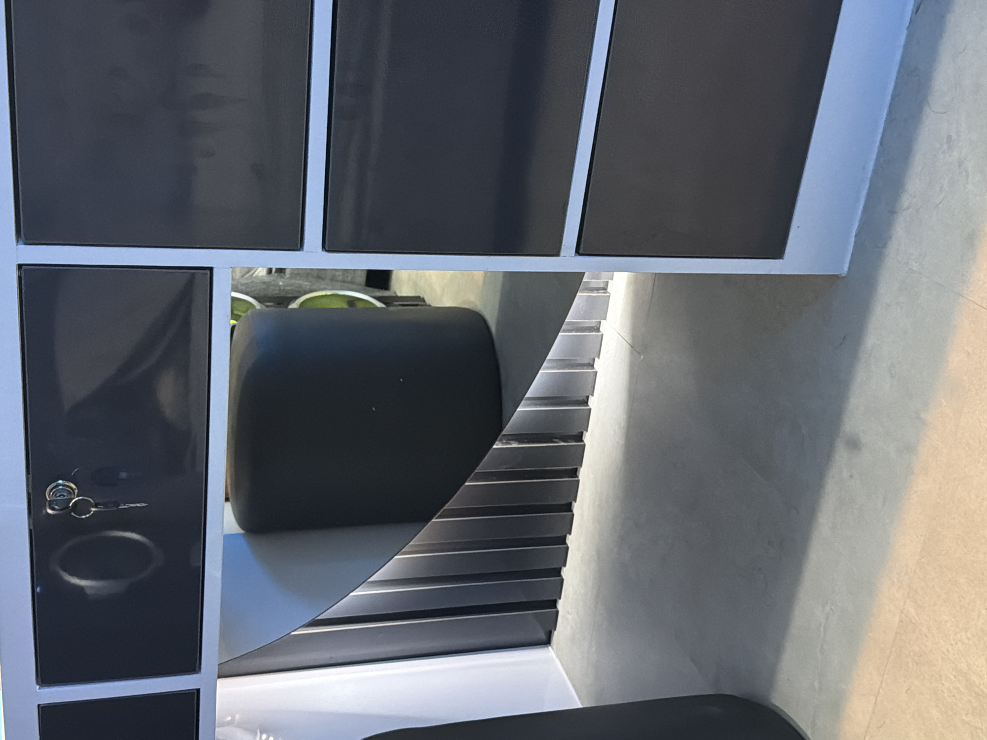 The image shows a close-up, eye-level view of a dark grey cabinet with light blue trim. The top portion of the cabinet features a drawer with a silver lock and two keys. The drawer is slightly ajar, revealing a circular indentation within it. Below the drawer, the cabinet has a curved opening that reveals a dark grey, ribbed structure. The floor is concrete and reflects light. The background shows more sections of the dark grey cabinet, suggesting it's part of a larger furniture unit. The scene is indoors.