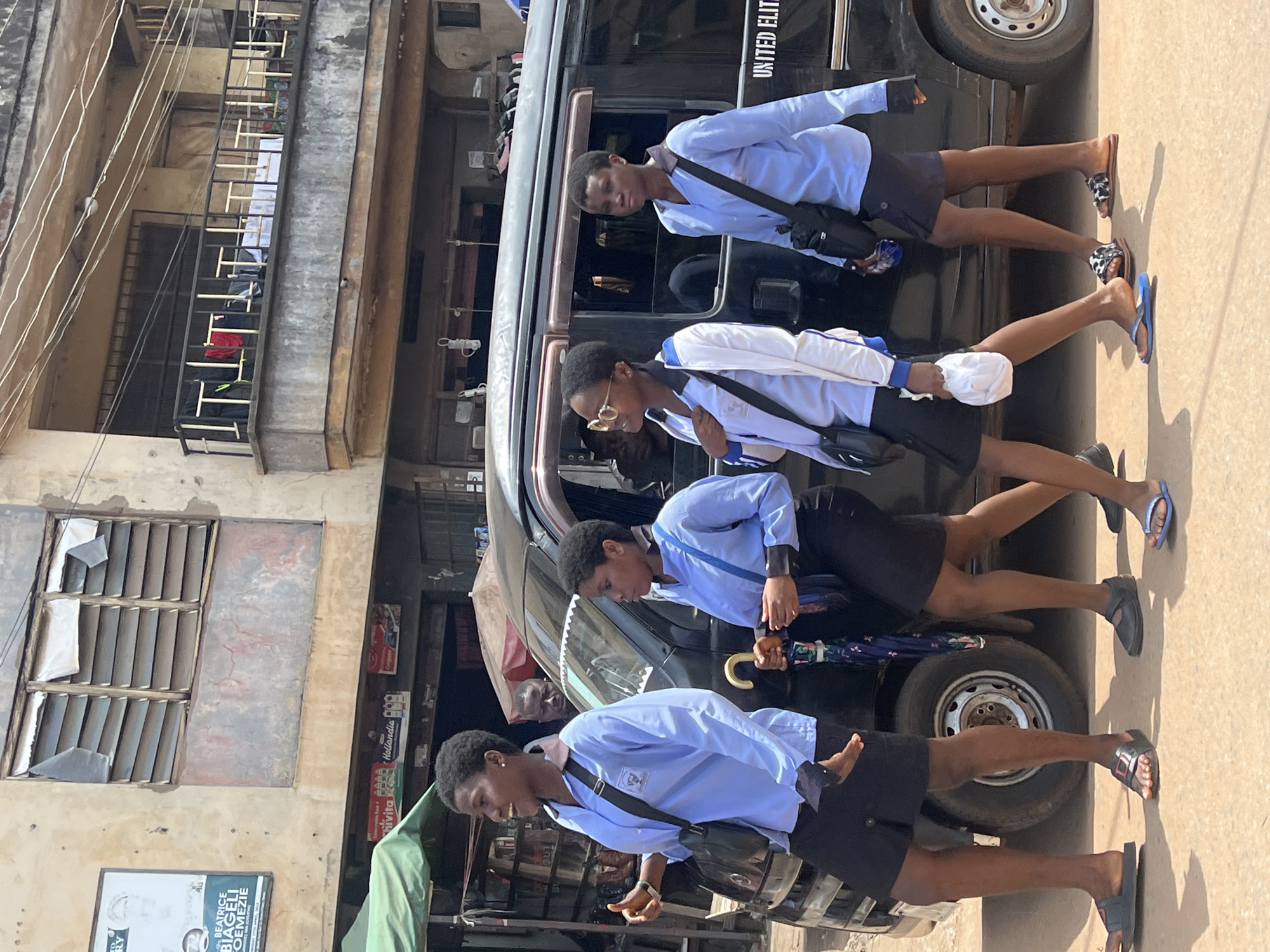 A group of four young individuals, appearing to be students, walk alongside a black van. They are dressed in light blue uniforms, suggesting a school setting. The individuals are seen from the waist down, showcasing their legs and footwear, and from the shoulders up, displaying their faces and torsos. Two of them are carrying bags, and one is holding a patterned umbrella with a golden handle. The scene is outdoors, with a paved surface that appears to be a road or sidewalk. Buildings are visible in the background, including a weathered structure with louvred windows. The lighting suggests it is daytime, possibly mid-morning or afternoon, with clear skies. The overall mood is casual, with the individuals seemingly engaged in a mundane activity like walking or commuting. The text "UNITED ELITE" is visible on the side of the van. There is also a sign that reads "BEATRICE BIAGELI OEMEIZIE" on a building in the background.