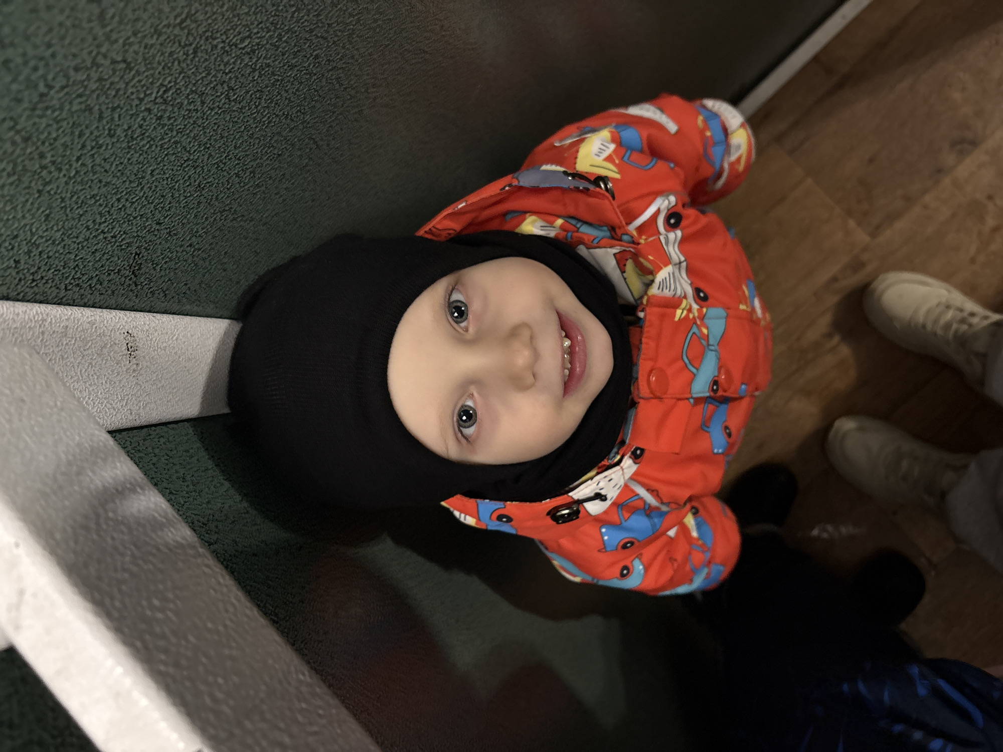 A young child wearing a black balaclava and a red and blue patterned winter jacket is looking up and smiling. They are seated on a padded bench in what appears to be a public transportation setting, possibly a bus or train. The bench is covered in a dark green textured material. The flooring is wood or a wood-like material. Nearby are white shoes belonging to someone likely accompanying the child. The location is Chaikovsky, Russia.