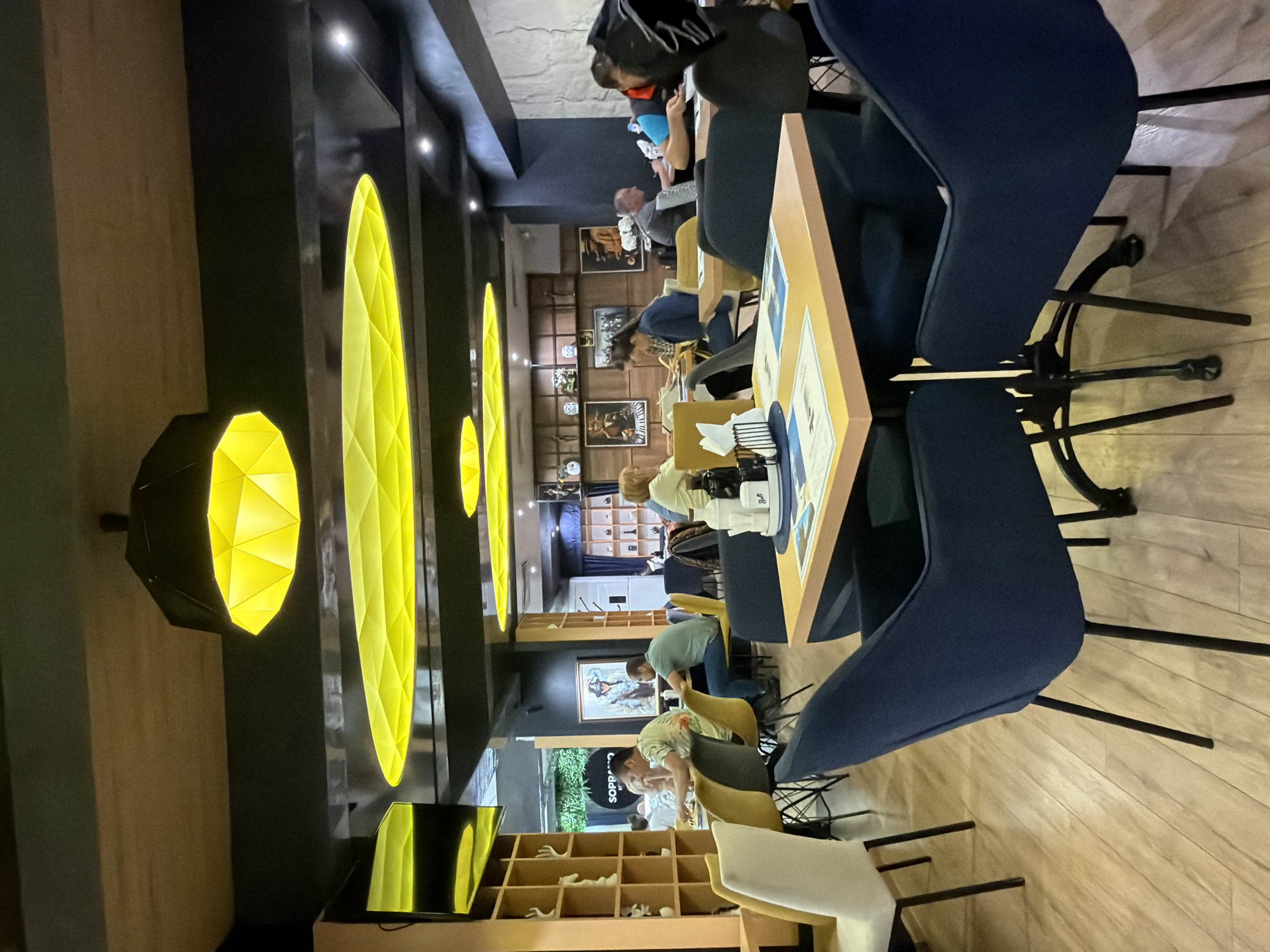The image captures the interior of a modern restaurant or cafe in Gabrovo, Bulgaria, filled with patrons. The scene depicts a contemporary design with dark walls and ceiling, contrasting with light wooden flooring and shelving units.
A defining feature of the decor is the striking yellow geometric lighting. Long, elongated yellow fixtures run along the ceiling, and several hexagonal or octagonal pendants hang lower, casting a warm glow. Additional white downlights provide ambient illumination.
Several customers are seated at rectangular tables with light wooden tops. The seating includes stylish dark blue chairs with black metal frames, as well as some yellow and white chairs with wooden legs. People are casually dressed, engaged in dining or conversation. Some individuals are seen looking down at their tables or phones. In the foreground, a person wearing a black hoodie is visible, while others are seated further back.
The walls are decorated with framed artwork or pictures. Wooden shelves display various decorative items, including abstract white sculptures. On the tables, condiment holders, napkin dispensers, and menus are present. Partial text "GOLF" is visible on a table item, and a sign on a wall or shelf reads "SOPRANO," likely indicating the establishment's name. The atmosphere appears relaxed and inviting, suggesting an evening or indoor setting given the reliance on artificial lighting.