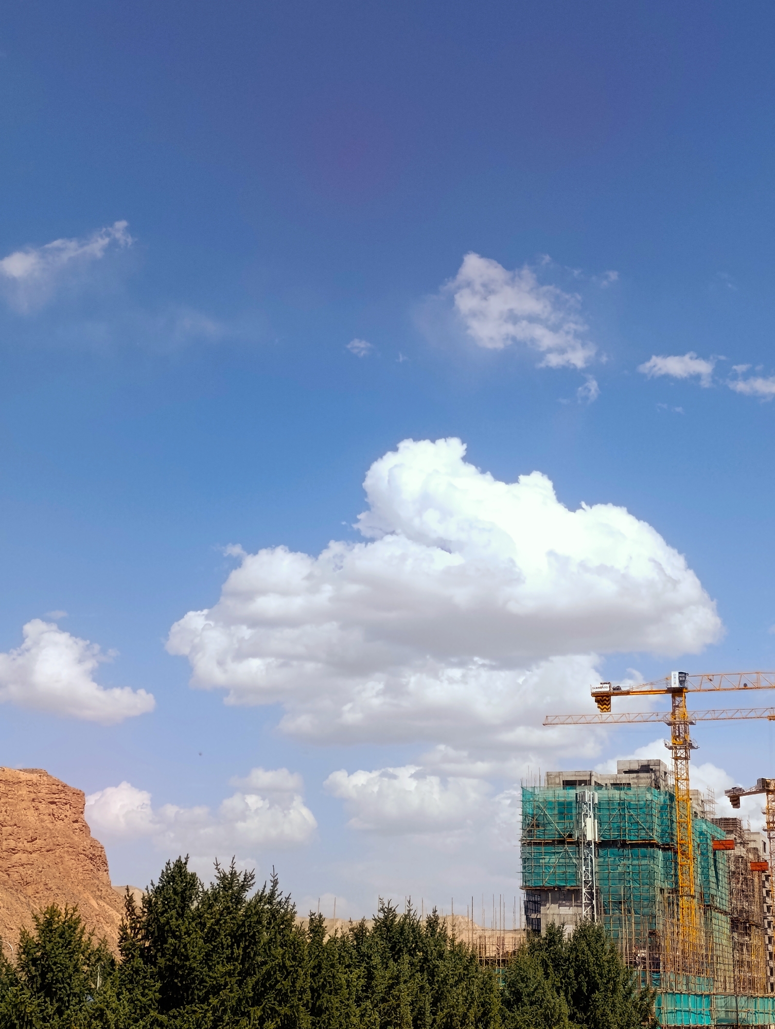 This is a photograph taken outdoors in the city of Naryn City, Kyrgyzstan, under a blue sky with scattered white clouds. The image captures a construction site in progress, with several tall, modern buildings partially obscured by scaffolding and green netting. Two large yellow construction cranes are prominently visible, extending over the buildings. In the foreground, a line of dark green coniferous trees forms a dense barrier across the bottom of the frame. To the left, a large, reddish-brown rock formation rises sharply from the landscape. The overall scene suggests urban development and the ongoing construction of infrastructure in a natural setting. There are no people visible in the image.