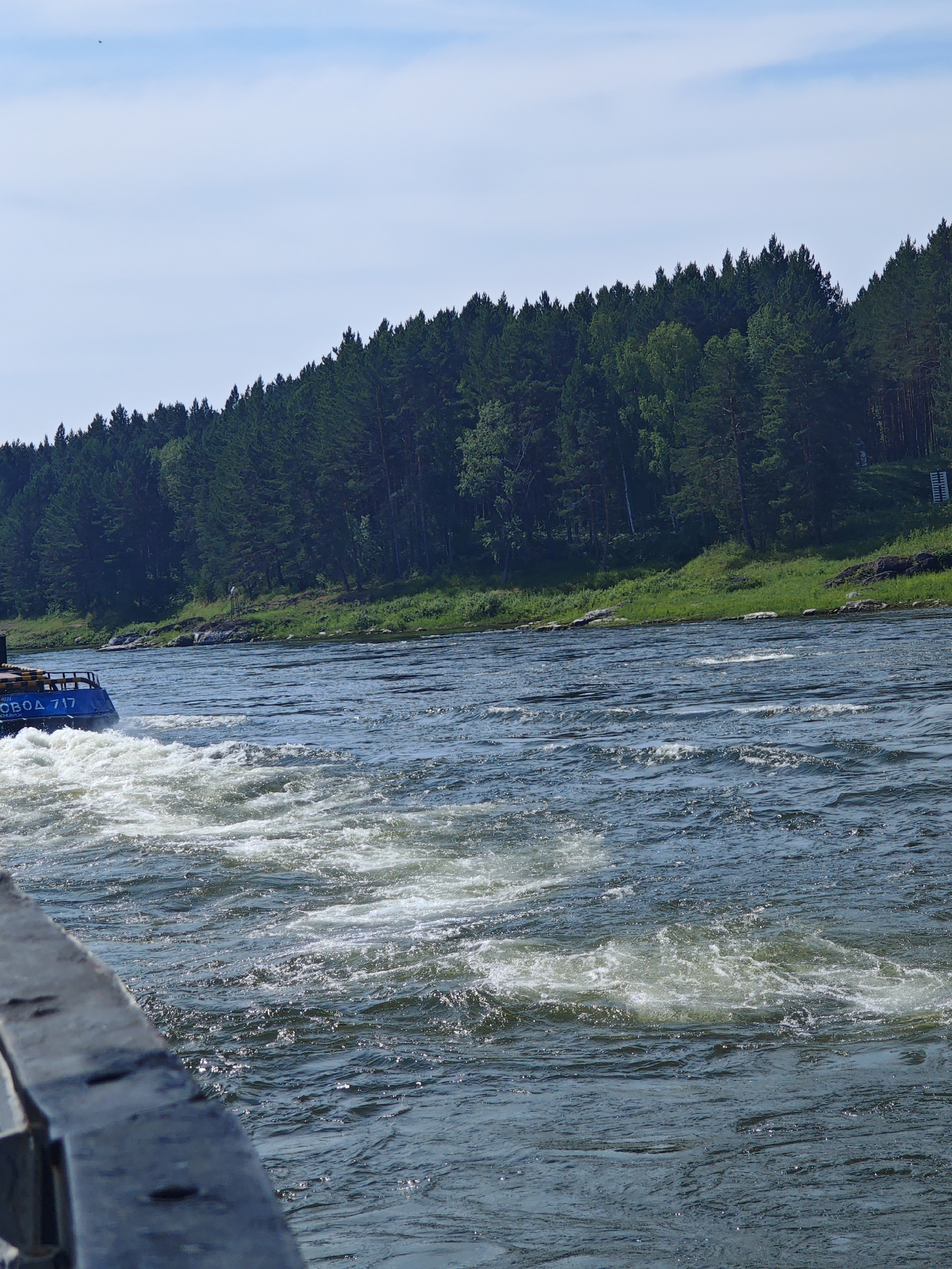 The image depicts a wide, turbulent river with a densely forested bank on the opposite side. In the foreground, a dark, weathered railing runs along the bottom edge of the frame, partially obscuring the view of the water. A blue boat, identified by Cyrillic lettering that reads "ВОДН0 717" (VodnO 717), is visible on the left side of the image, creating a wake of white foam as it moves through the water. The river itself is a deep blue, with ripples and churning white water indicating a strong current. The forest on the far bank consists of numerous evergreen trees, densely packed together, with a strip of green grass at the water's edge. The sky above is a clear blue with a few wispy white clouds. The scene appears to be a natural landscape with a functional boat operating on a river. The location is identified as Porog, Russia.