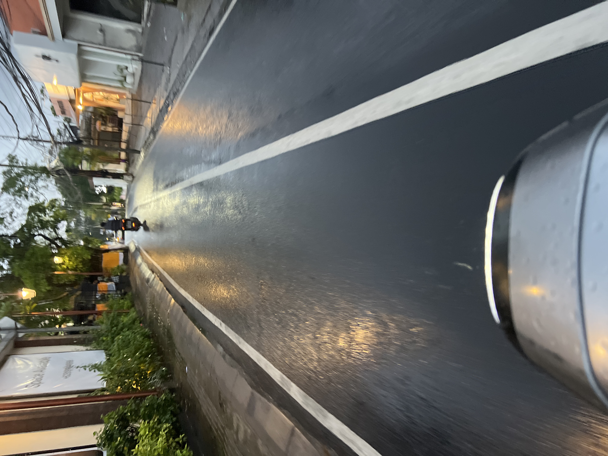 A low-angle, POV shot captures a wet road in North Kuta, Indonesia, likely during dusk or early morning given the overcast sky and artificial lighting. The asphalt surface is slick with rain, reflecting the warm glow of street lamps and building lights, creating streaks of light across the dark road. A white line marks the center lane and another runs along the edge of the sidewalk. A single scooter, ridden by a person wearing black clothing, is visible in the distance, also traveling on the wet road. On the left side of the street, a building with large banners indicating "Space Available" is partially obscured by lush green foliage. The architecture of the buildings on the right suggests a commercial or residential area. The scene conveys a sense of normalcy in an urban environment, with everyday activities continuing despite the wet conditions.