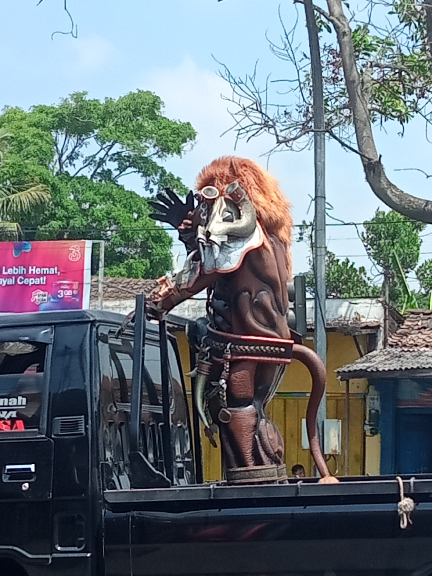 A dark-colored pickup truck is loaded with a large, brown, demonic-like effigy in the city of Urutsewu, Indonesia. The effigy has a furry orange mane, a menacing mask with goggles, and outstretched black gloves. It is adorned with decorative belts and what appears to be a horn or tusk. The truck's bed is filled with the effigy, which stands tall, obscuring the background. In the background, there are trees, a utility pole, some buildings with tiled roofs, and a billboard with Indonesian text. The sky is clear and blue.