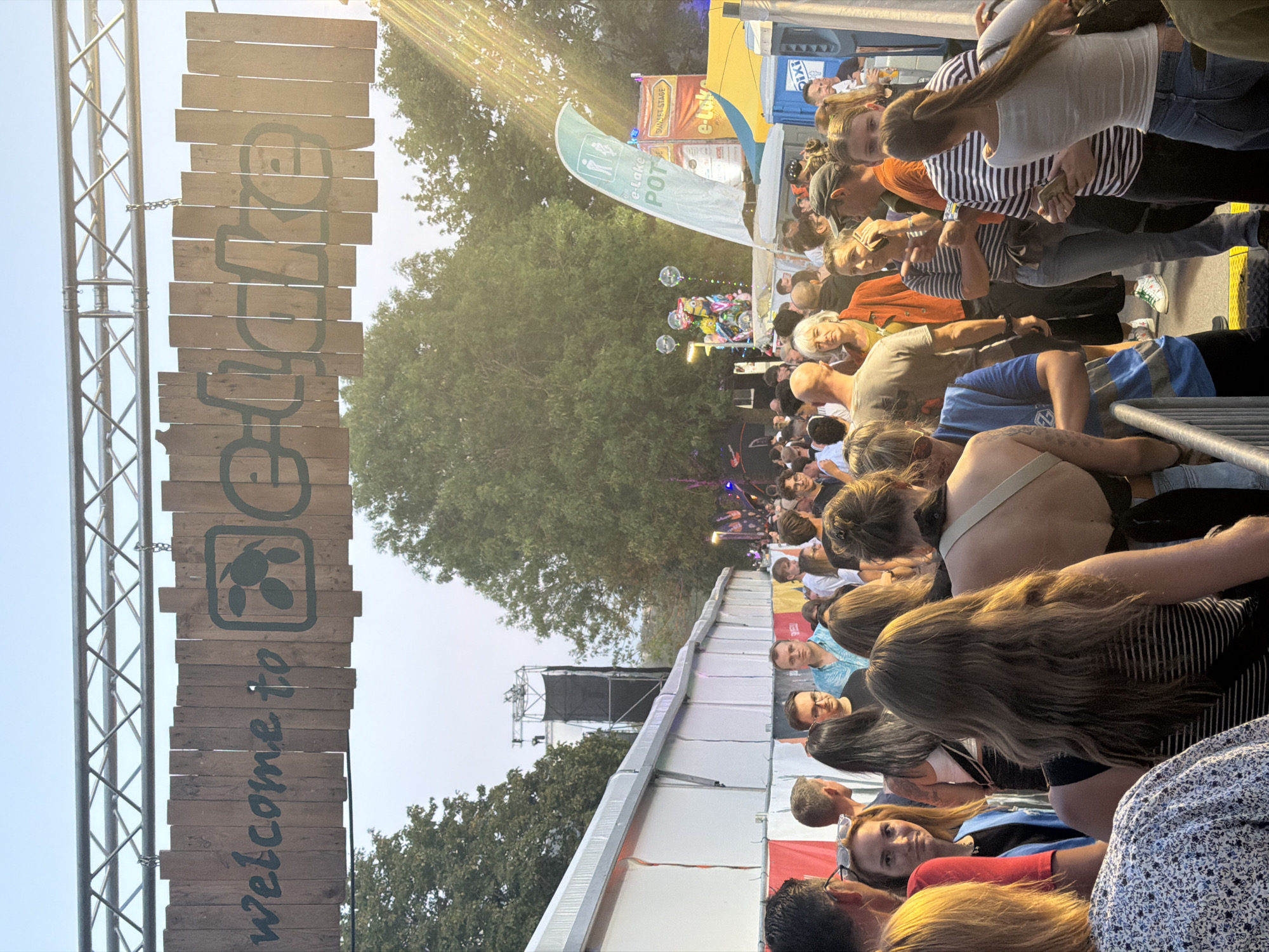 The image captures an outdoor event, likely a festival, at the e-Lake festival grounds in Echternach, Luxembourg. A wooden sign with "welcome to e-Lake" is prominently displayed, hanging from a metal structure. In the foreground, a crowd of people are gathered, facing towards the event area. The crowd is diverse in age and gender, with many individuals dressed in casual attire. Some people are engaged in conversation, while others are looking around or moving through the space.
Behind the crowd, white tents line the perimeter of the festival grounds. In the background, trees and a stage structure are visible. A blue and white flag with pictograms of a toilet and shower is visible, along with signs indicating "SUNSET STAGE" and "e-Lake POT". There are also balloon decorations featuring characters from the movie "Despicable Me". The lighting suggests it is either late afternoon or early evening, with sunlight casting rays through the trees. The overall atmosphere appears to be one of a lively public gathering.