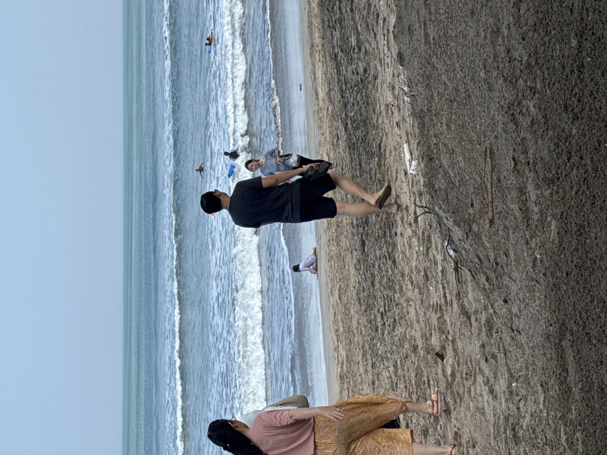 The image depicts a beach scene in Kuta, Indonesia, with the ocean and sky dominating the background. In the foreground, the sandy beach is visible, with some debris and footprints. Several people are present on the beach and in the water.
On the left side of the image, a woman with dark hair is walking away from the camera, wearing a pink top and patterned pants. A person, partially obscured, stands next to her. Further inland, a child in a colorful swimsuit is sitting on the sand.
In the middle ground, a man in a dark t-shirt and shorts is walking towards the ocean, carrying a shoe. Beside him, a woman in a gray top and dark pants is looking towards the ocean.
In the water, several individuals are visible engaged in water activities. Some appear to be surfing or using bodyboards, with their boards in the water. The ocean has moderate waves breaking near the shore. The sky is overcast, with a pale blue hue. The overall atmosphere suggests a leisurely day at the beach in a popular tourist location.