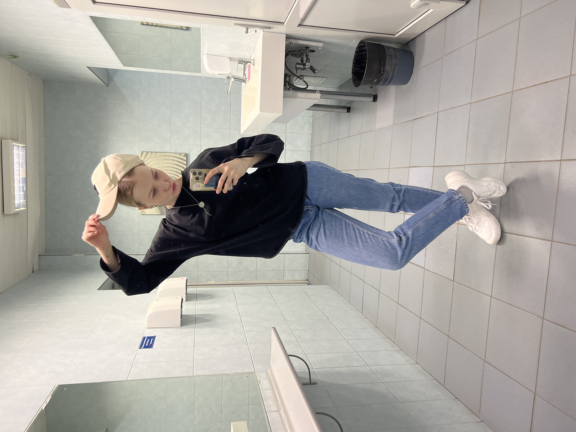 A full-body mirror selfie captures a young person in what appears to be a public restroom in Khimki, Russia. The person, likely female, has light hair and is wearing a beige baseball cap, a loose-fitting black long-sleeved top, light blue denim jeans, and white chunky sneakers with white socks. She wears a necklace with a round pendant and a visible earring.
The subject holds a smartphone with a decorative case in her right hand, pointing it towards the mirror to take the photo. Her left hand is casually holding the brim of her cap. She stands with her weight slightly shifted, her expression neutral.
The restroom features light blue/gray tiled walls and a light gray tiled floor. In the background, a white sink with chrome faucets is visible to the right, under which a black mesh waste bin is situated. Above the sink, a larger mirror reflects more of the room. To the left, a rectangular fluorescent light fixture is mounted on the wall. Two white hand dryers are visible on the wall behind the person.
A small blue sign with white symbols or text is affixed to the wall near the hand dryers, but its content is illegible. The phone case also displays small, indistinct patterns or text. The overall setting suggests a functional and well-lit public facility.