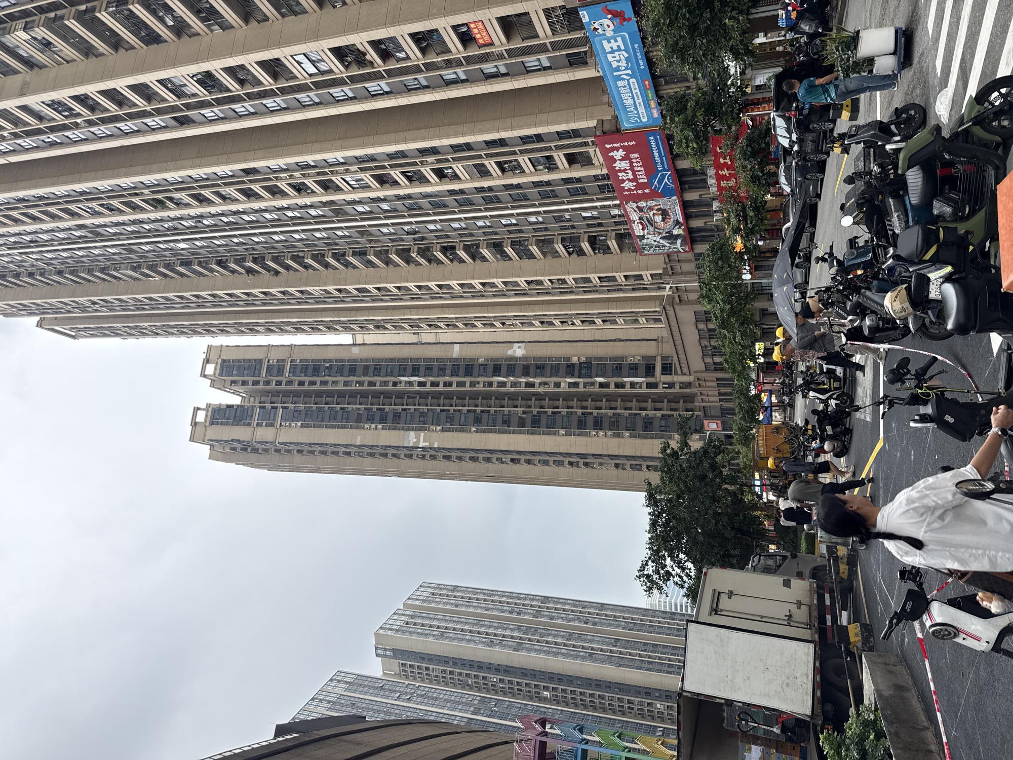 The image depicts a street scene in Haizhu District, China, with a focus on tall, modern residential buildings. In the foreground, a woman in a white shirt with her hair in a braid is walking away from the camera, and several electric scooters are parked on the street. Behind her, a white delivery truck with its rear door open is parked, with goods visible inside.
In the middle ground, a group of people, some wearing yellow hard hats, are gathered near the truck. A black umbrella is providing shade for some of them. Further down the street, more electric scooters are parked, and several cars are visible, including black SUVs. On the sidewalk, people are walking, and there are potted plants.
The background is dominated by several very tall apartment buildings with numerous windows and balconies. Billboards with Chinese text are displayed on the lower levels of some of these buildings. The sky is overcast and grey, suggesting a cloudy day. The environment appears to be an urban residential area with significant development.