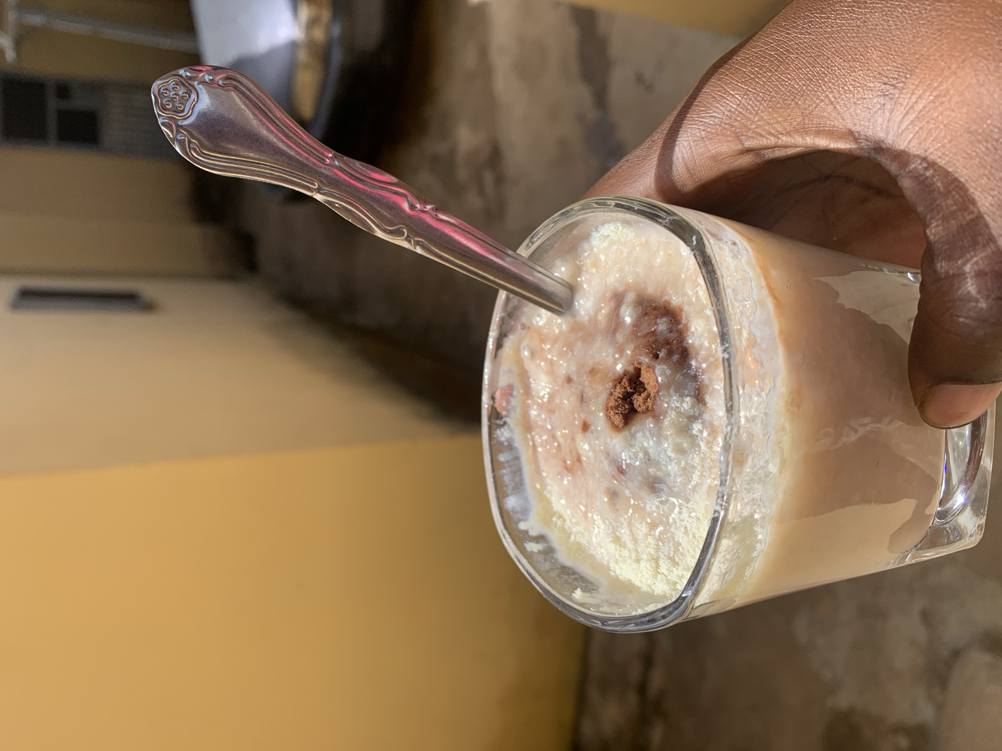 A close-up shot shows a person's dark-skinned hand holding a glass of a beverage. The drink appears to be a smoothie or shake, with a lighter-colored layer on top, sprinkled with dark brown powder. A silver spoon with an ornate handle rests in the glass. The background is a blurred outdoor setting, with a yellow wall on the left and a textured surface, possibly concrete, in the background. The location is Rafin Par, Nigeria.