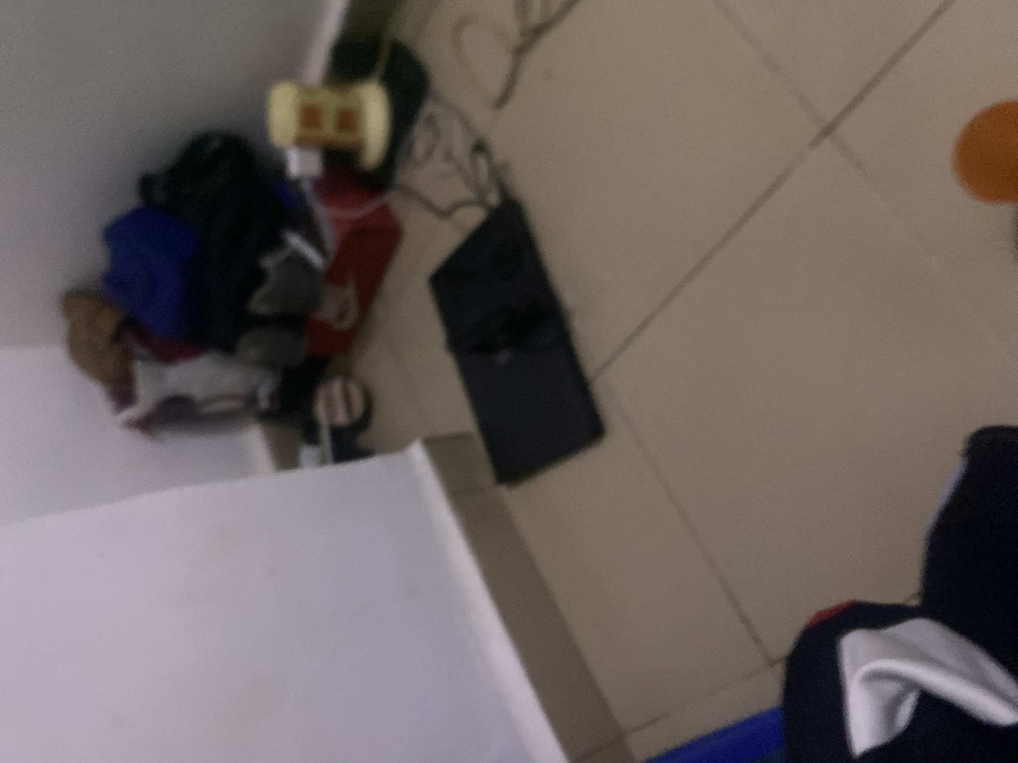 The image depicts a cluttered corner of a room. A pile of clothing, including a blue item and a tan-colored bag, is situated against a light-colored wall. To the right of the clothing pile, a yellow cylindrical object with orange markings sits on a black mat. In the foreground, a flat, black object, possibly a laptop or a tablet, rests on the tiled floor. The overall environment suggests a lived-in space with items scattered around. There is no information within the image to suggest a specific location in Howayi, Nigeria.