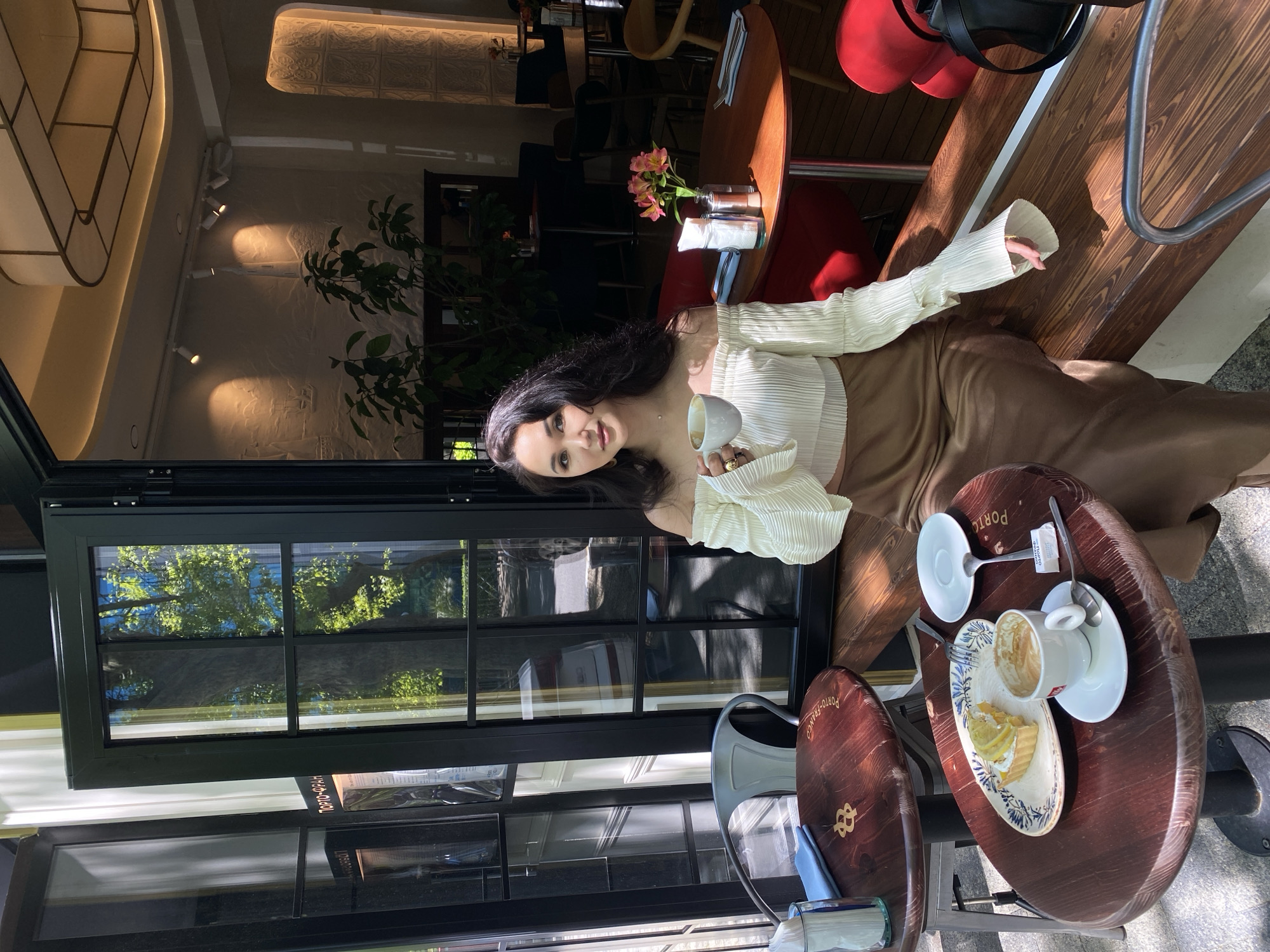 The image depicts a young woman seated at a wooden table inside a bright cafe or restaurant. She has long, dark hair and is wearing a white, off-shoulder pleated top and a brown, flowing skirt. She holds a white cup in her right hand, adorned with a gold-colored ring, and looks towards the camera with a gentle smile.
The setting is illuminated by natural sunlight streaming through large, dark-framed windows to the left, casting distinct shadows on the light-colored floor and textured walls. Beyond the windows, reflections of lush green trees and a blue sky indicate a sunny outdoor environment. The cafe's interior features modern track lighting, a decorative ceiling panel, and plants. In the background, other dark wooden tables and red upholstered chairs are visible.
On the table in the foreground, directly in front of the woman, sits a white plate holding a slice of lemon tart or cake and a fork. An empty white coffee cup with a saucer and a sugar sachet are placed nearby. Another empty cup and saucer are also on the table. The dark wooden table surface is branded with the text "PORTO-FRANCO" and a stylized logo, resembling an anchor or an '8'.
The activity taking place is a relaxed cafe visit, where the woman is enjoying a beverage and dessert. She appears to be posing for the photograph. The overall atmosphere is bright, warm, and inviting, suggesting a pleasant daytime experience in a cafe, fitting the context of Sochi, Russia.