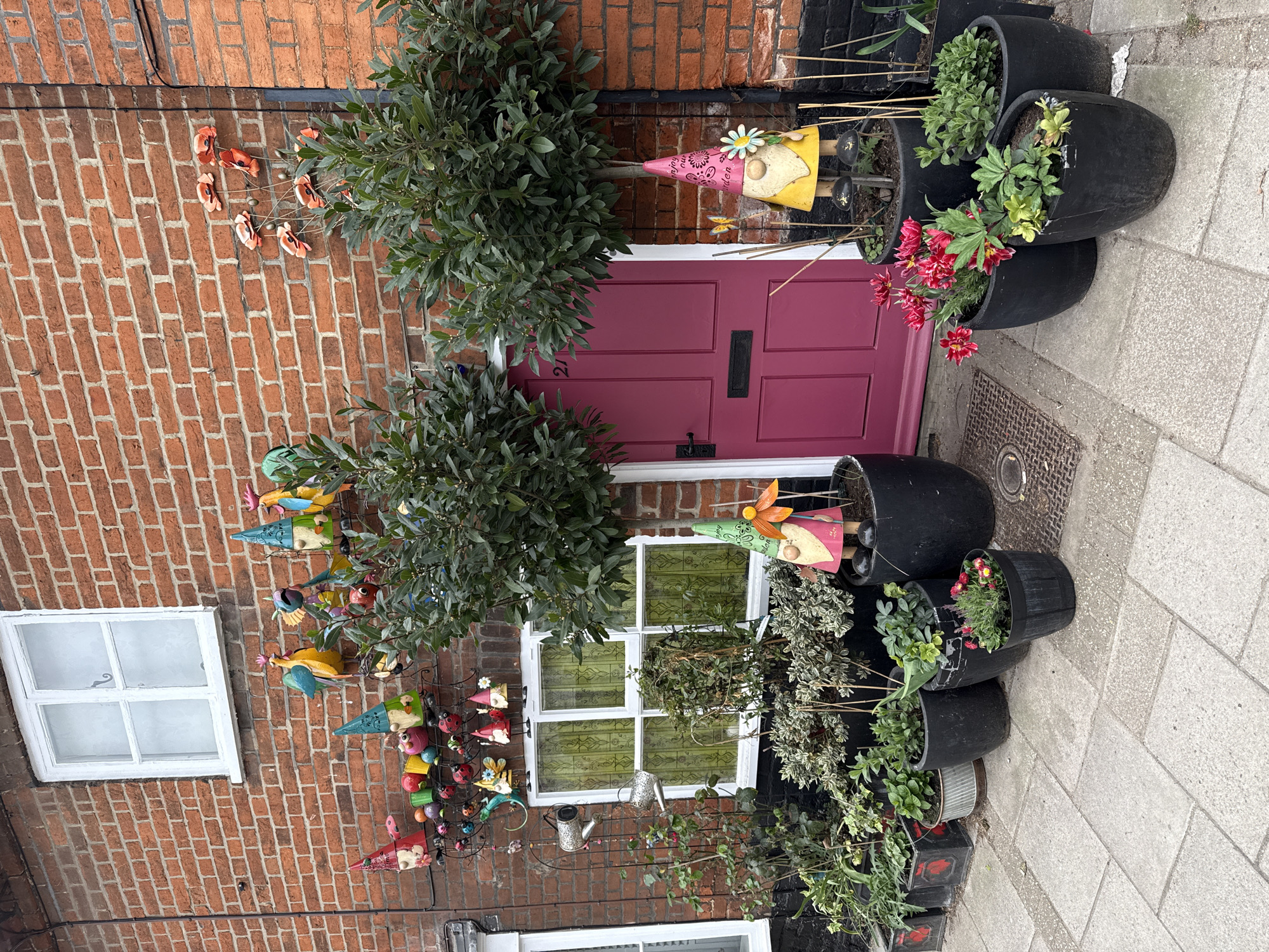 Exterior view of a two-story brick building in a city within East Suffolk, United Kingdom. The main focus is a pink front door with a black letter slot. A white-framed window is located to the left of the door and another below it. The walls are adorned with decorative metal art pieces depicting birds, gnomes, and garden-themed figures. Trees and potted plants decorate the front entrance. The sidewalk is paved with light gray slabs. There are no people visible in the image.