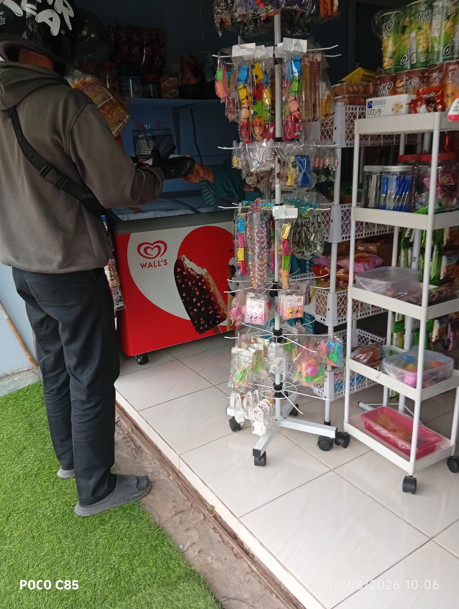 A person wearing a gray hoodie and black pants stands in front of a Wall's ice cream freezer and a display rack of toys and snacks. The person is wearing a black helmet with white graphics and has a strap from a bag across their chest. The Wall's freezer has a prominent red and white design with an image of an ice cream cone. To the right of the freezer is a white multi-tiered display rack filled with various items, including colorful toys, small packaged snacks, and what appear to be pens or straws in plastic containers on the top shelves. The floor is tiled, with a strip of artificial green turf to the left. The scene appears to be an outdoor market stall or a small convenience store in South Tangerang, Indonesia, indicated by the text watermark "POCO C85" and the date "12/08/2026 10:06" which are typical of a mobile phone timestamp.