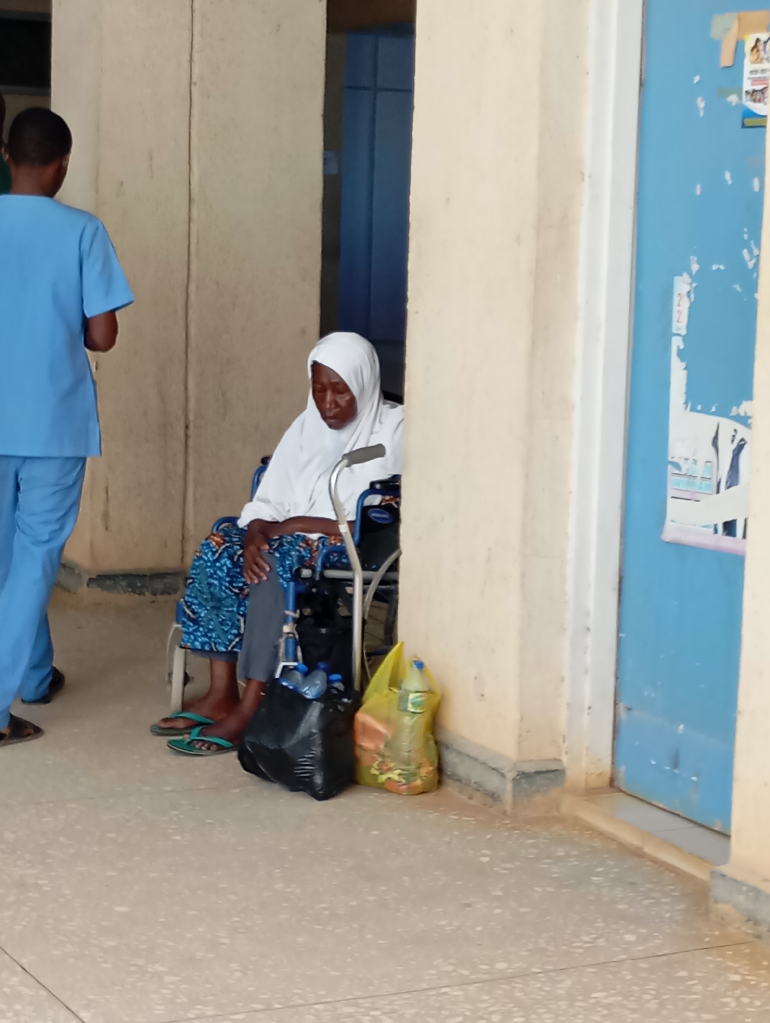 In Gwong, Nigeria, an elderly woman in a white hijab sits in a wheelchair in a hospital waiting area. She is wearing a blue patterned dress and gray pants. Next to her are a black plastic bag containing water bottles and a yellow plastic bag. A medical professional in blue scrubs walks away from her. The scene takes place in a hallway with cream-colored walls and a blue door.