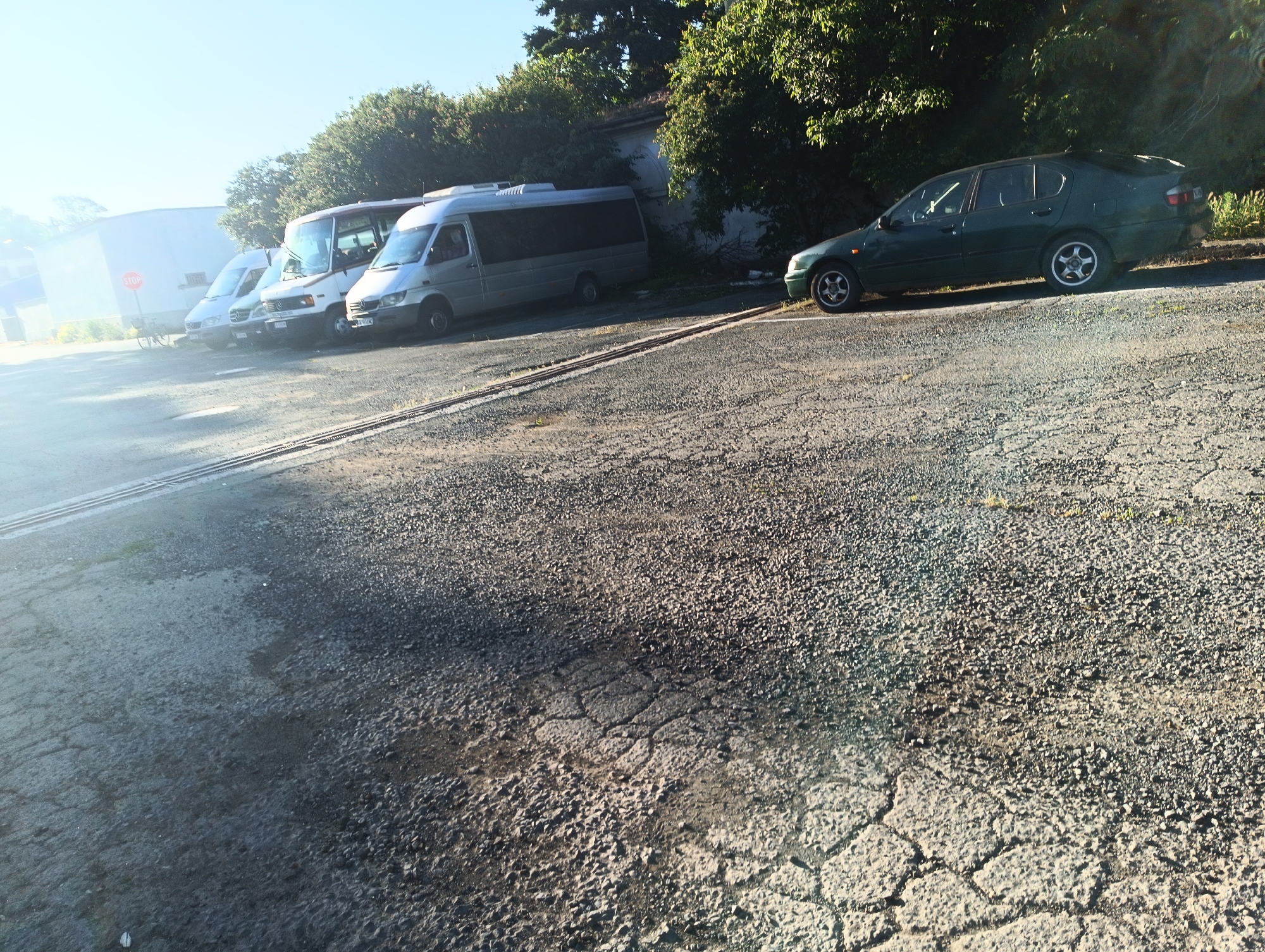 The image captures a sunny outdoor scene, likely a parking area, with several vehicles present. In the foreground, a cracked asphalt surface dominates the lower half of the frame. Behind the cracked surface, a line of vehicles is parked. Closest to the viewer, a green sedan is parked on the right side. To the left of the sedan, a silver minibus is parked. Further to the left, two more white vans and a larger bus are visible, all parked in a row. In the background, lush green trees and foliage line the scene, with a light-colored building visible behind the vans. The sky is a clear, bright blue, suggesting fair weather. The overall impression is of a quiet, possibly industrial or commercial area. This scene is located in Karnobat, Bulgaria.