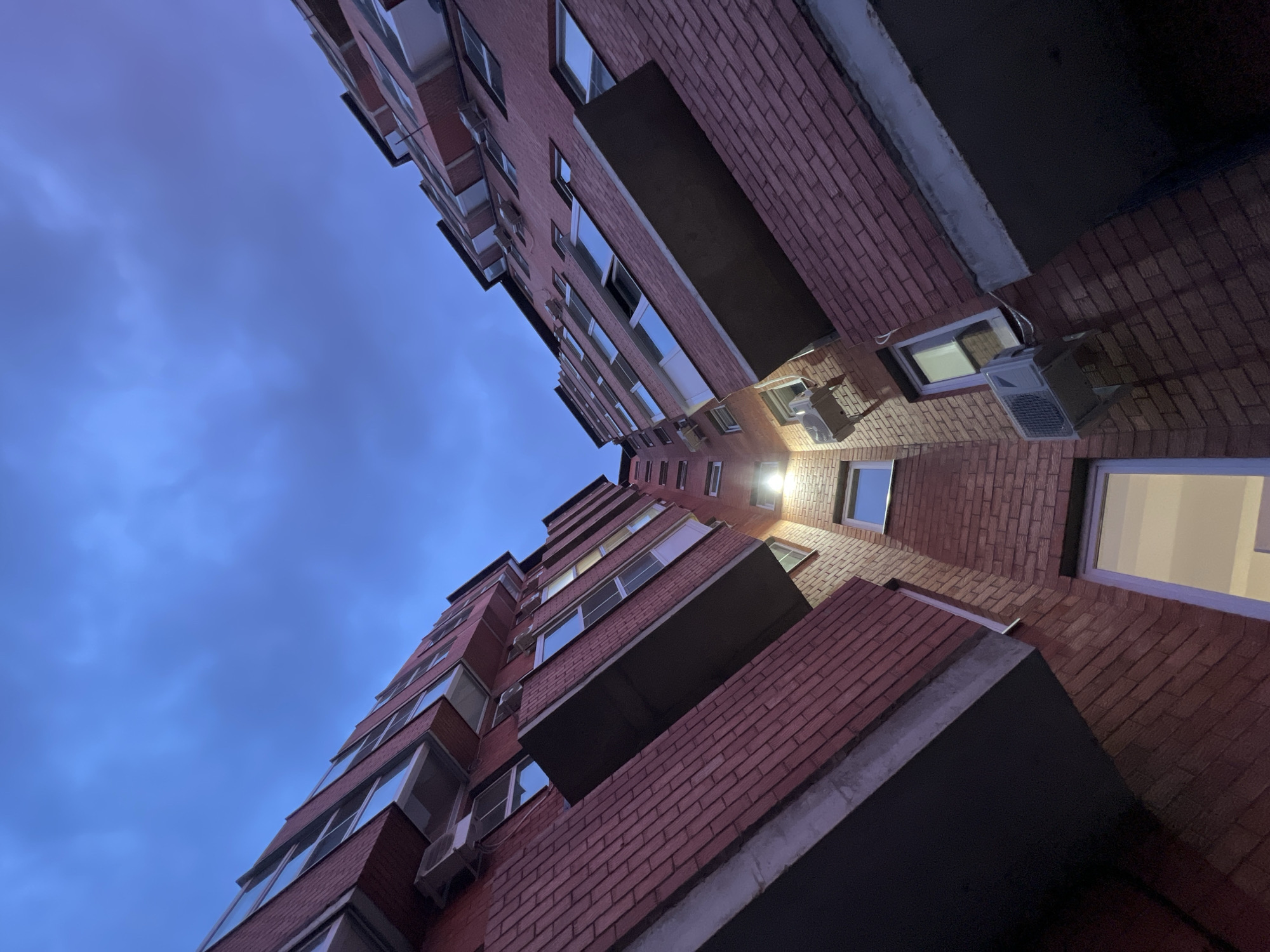 The image is a low-angle shot of two brick apartment buildings in Yablonovskiy, Russia. The buildings are facing each other, with a narrow strip of sky visible between them. The sky is a deep blue, indicating either dusk or dawn. The buildings are multi-story, with balconies and windows visible on each floor. Many windows have white air conditioning units attached to the exterior. A bright light shines from one of the buildings, illuminating the brickwork. The perspective emphasizes the height of the buildings and the close proximity between them.