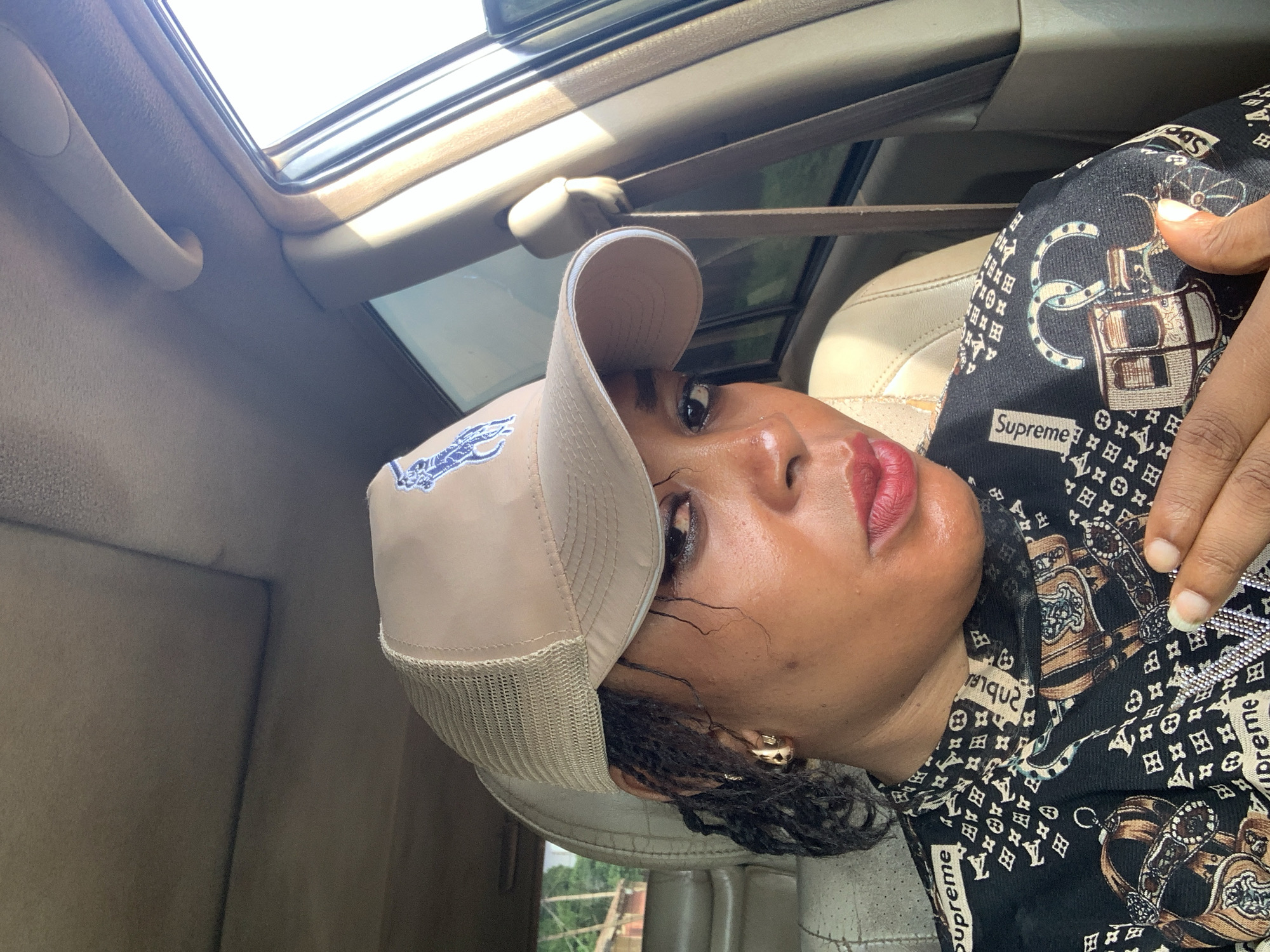 The media file, presented rotated 90 degrees counter-clockwise, depicts a person from the chest up, seated inside a car during what appears to be a sunny day in Sabon Tasha, Nigeria. The person is looking directly at the camera with a neutral expression, wearing red lipstick.
They are wearing a beige baseball cap with a mesh back, featuring a blue embroidered logo resembling a polo player. Their dark, braided hair is visible beneath the cap, and a small gold earring adorns their right ear. The person is dressed in a black top patterned with various motifs including chains, stylized horse-drawn carriages, and repeated small square monograms. The word "Supreme" is distinctly visible printed on the fabric of the top in several places. A hand with a light-colored fingernail rests on the person's right shoulder, partially visible.
The car's interior is discernible in the background, featuring a light-colored headliner, an overhead grab handle, and a sunroof, through which bright daylight pours in. Elements of a beige car seat and a safety belt are also visible, confirming the setting as a vehicle interior. The strong natural light indicates a clear, sunny weather condition. The image captures a moment of the person posing inside the car.
