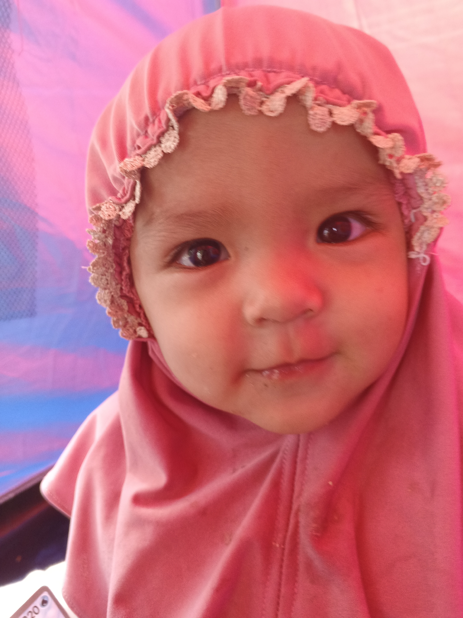 A close-up, head and shoulders shot shows a young child wearing a pink hijab with a lace trim. The child's face is turned towards the camera, with their eyes looking slightly to the side. The background is a blurred mix of pink and blue, possibly indicating a tent or fabric backdrop. A small portion of a phone screen with the text "20" is visible in the lower left corner of the frame. The scene appears to be indoors or in a covered outdoor space. The location is identified as Perbawati, Indonesia.