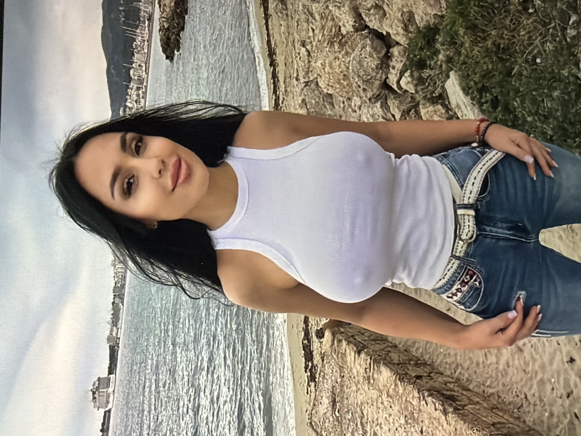 The image shows a woman standing on a rocky beach with the sea in the background. She has long, dark hair and is wearing a white tank top and blue jeans. Her hair is blowing in the wind, and she is looking at the camera with a slight smile. The sea is calm, and there are a few boats in the distance. The sky is cloudy. There is no indication of the city of Shumen, Bulgaria, in the image.