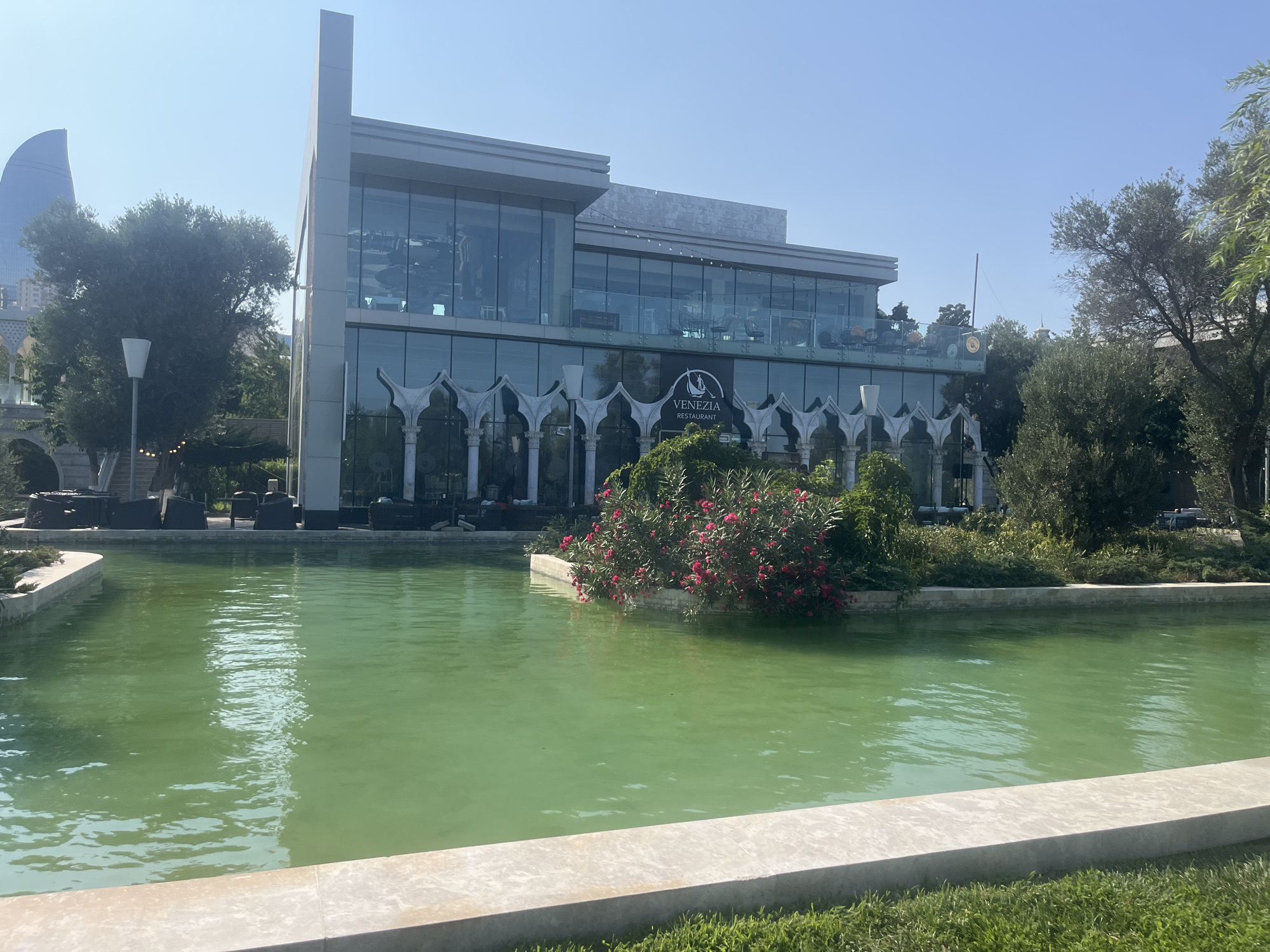 The image shows a view of the Venezia Restaurant in Sabail Raion, Azerbaijan, on a sunny day. In the foreground, a bright green pond reflects the clear blue sky and surrounding greenery. The pond is bordered by a light-colored concrete edge, with grass growing in front of it. Behind the pond, a modern building with large glass windows and a light-colored facade stands prominently. The building has multiple levels, with seating areas visible on the upper floors. Below the glass, there are decorative arches with white pillars. A sign with the text "VENEZIA RESTAURANT" is visible on the building. Lush green trees and flowering bushes, with bright pink flowers, are situated around the building and in the pond area. In the background, a distinctive flame-shaped skyscraper, the Flame Towers, can be seen in the distance, identifying the location as Baku, the capital of Azerbaijan, and specifically Sabail Raion. No people are clearly visible in the image. The overall scene depicts a well-maintained outdoor dining and recreational area.