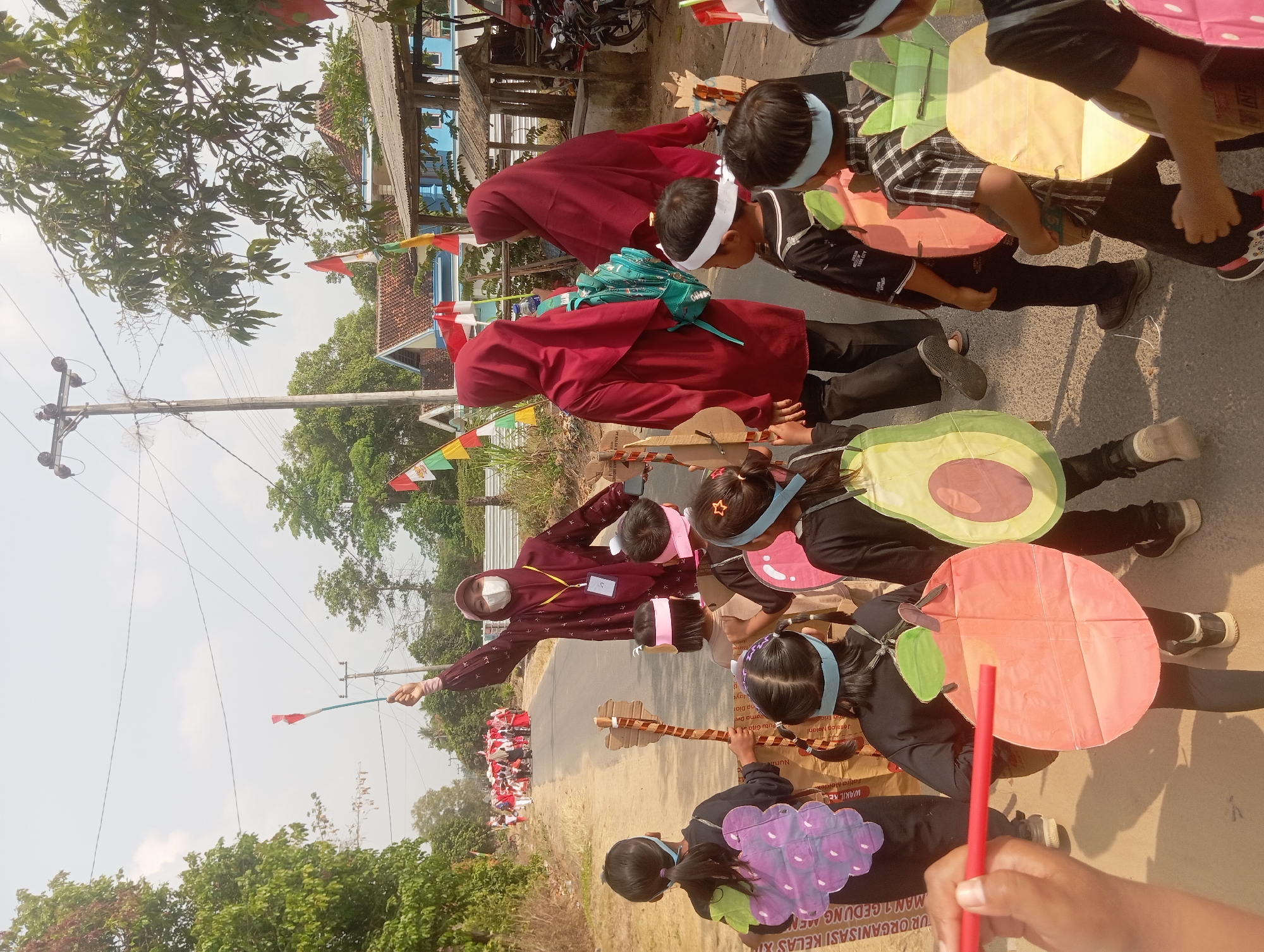 A group of children and adults are participating in a parade on a paved road in Dente Teladas, Indonesia. The children are dressed in costumes representing fruits, with some wearing apple, grape, and avocado cutouts. One child holds a red object, possibly a toy. An adult in a maroon hijab and mask is holding a stick with red and white flags. Other adults in maroon hijabs are also present, with one wearing a turquoise backpack. The parade is taking place on a sunny day, with trees lining the road and houses visible in the background. Flags are visible on poles and buildings, suggesting a festive occasion. The overall scene depicts a community event or celebration.