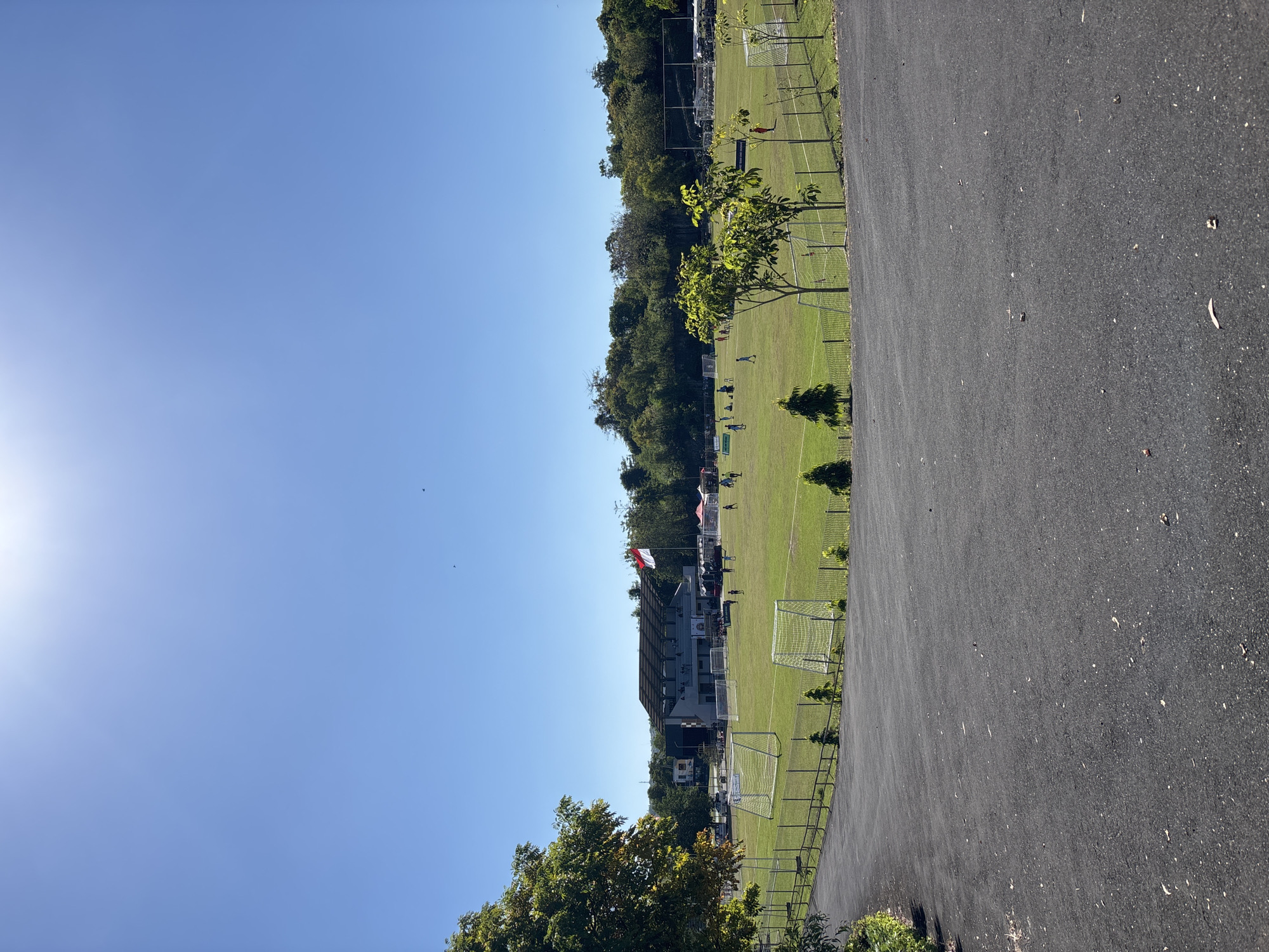 The image depicts a soccer field in Kuta Selatan, Indonesia. The field is green and well-maintained, with soccer goals at each end. There are people playing soccer on the field, and spectators are gathered around the perimeter. The surrounding environment includes trees, buildings, and a paved area. The sky is clear and blue. A red and white flag flies from a building near one end of the field.