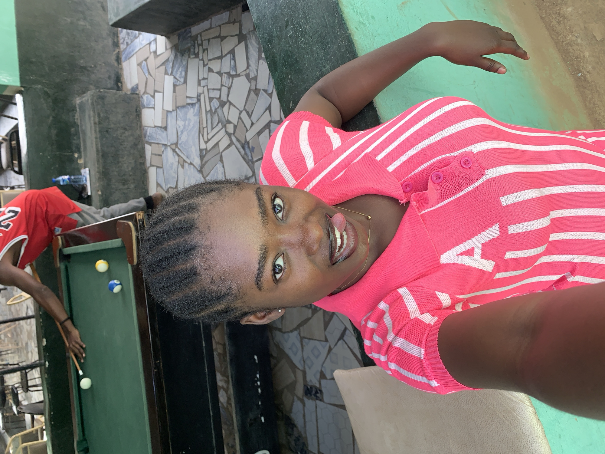 A young woman is taking a selfie in what appears to be a game room in Kabong, Nigeria. She is wearing a pink and white striped top, has her hair braided, and is sticking her tongue out. In the background, a person wearing a red jersey is playing pool. There is a pool table with three visible balls (white, yellow, and blue). The room has mosaic-tiled walls and stairs nearby.
