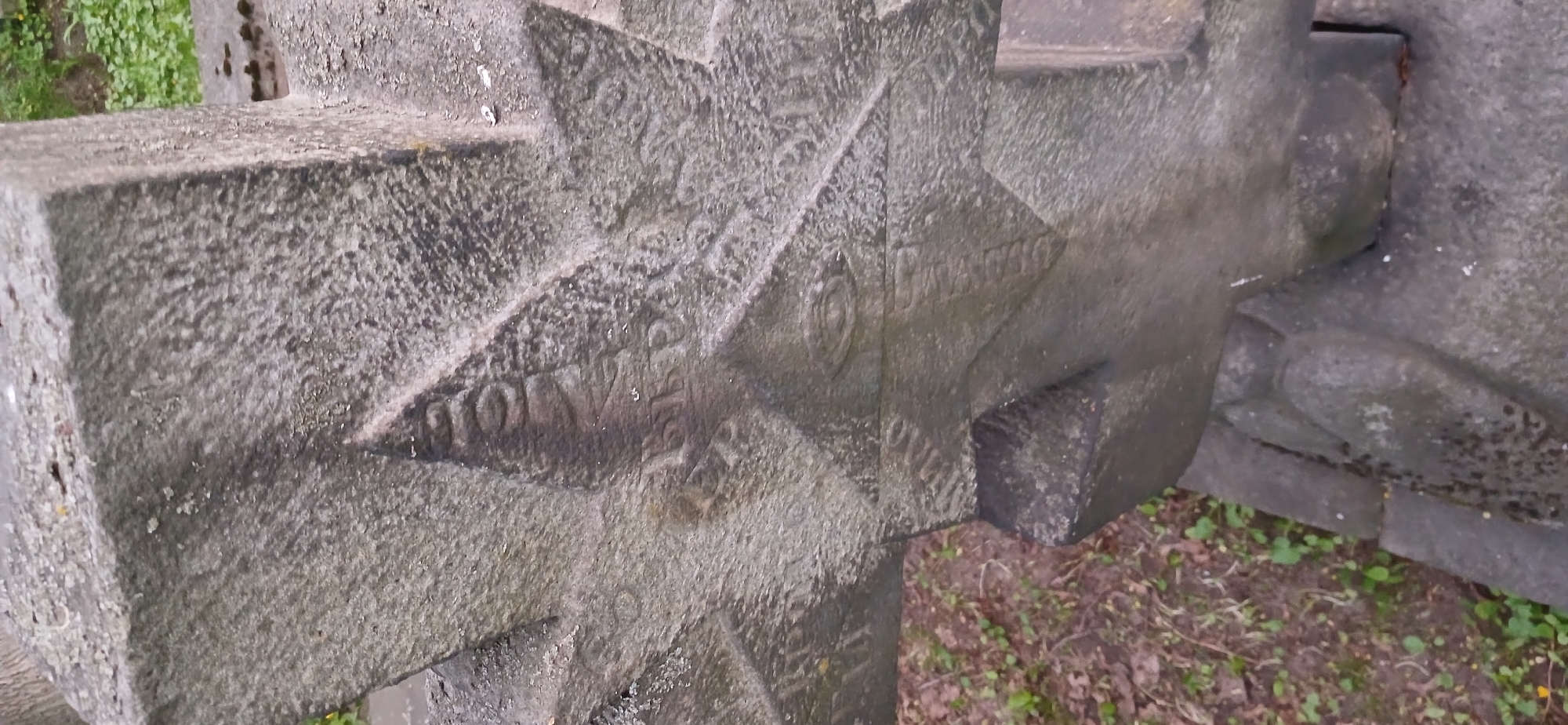 The image shows a close-up of a weathered gray stone cross, possibly a monument or grave marker. Inscriptions in Cyrillic are visible on the surface. The inscription includes the Cyrillic word "DOVOJNA" and the inscription is stamped with the "OTAKKO" logo. The cross is located outdoors, as indicated by the presence of soil and sparse vegetation in the surrounding area. Based on the logo and lettering on the cross, it is identified as being in Moscow, Russia.