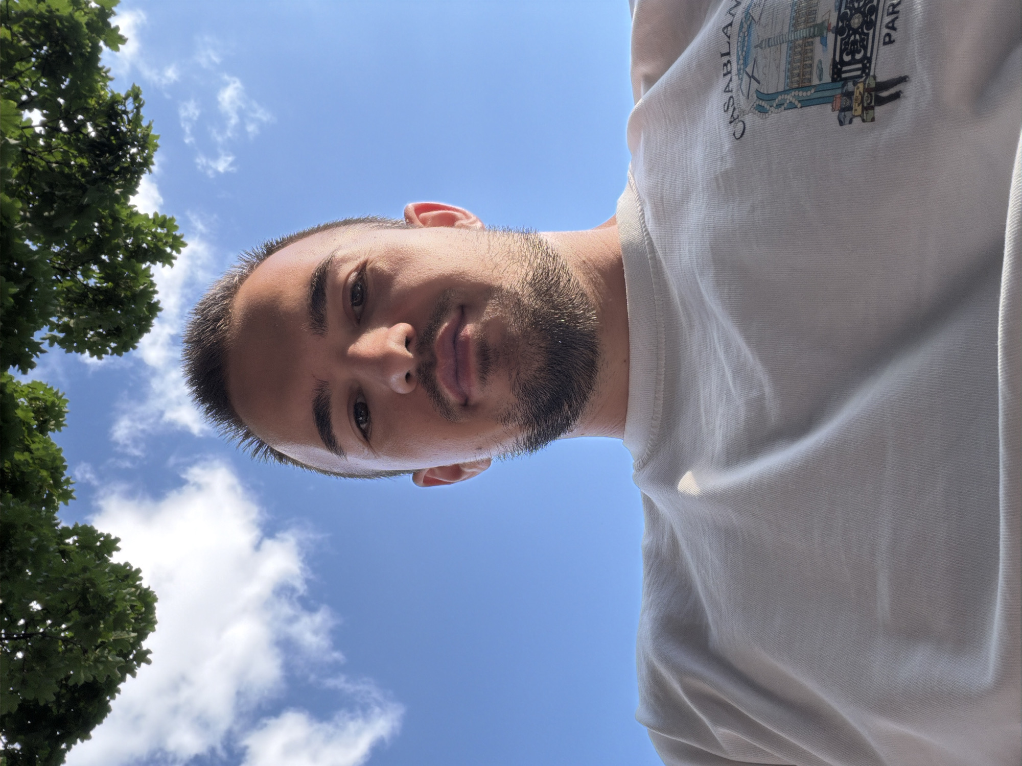 A low-angle, upward-facing photograph captures a young adult Caucasian male from the chest up, positioned against a clear blue sky with scattered white cumulus clouds. Green tree foliage is partially visible in the upper left and right corners of the frame. The subject has short dark hair, well-defined eyebrows, and a neatly trimmed dark beard and mustache. He looks directly at the camera with a neutral expression. He is wearing a light-colored, possibly white or cream-colored, short-sleeved crew-neck t-shirt. On the right side of his chest, a graphic design featuring architectural elements and text, including "CASABLANCA" and "PARIS," is partially visible. The lighting indicates an outdoor daytime setting with direct sunlight. The specific location within the city of Sofia, Bulgaria, cannot be determined from the visual information present in the image.