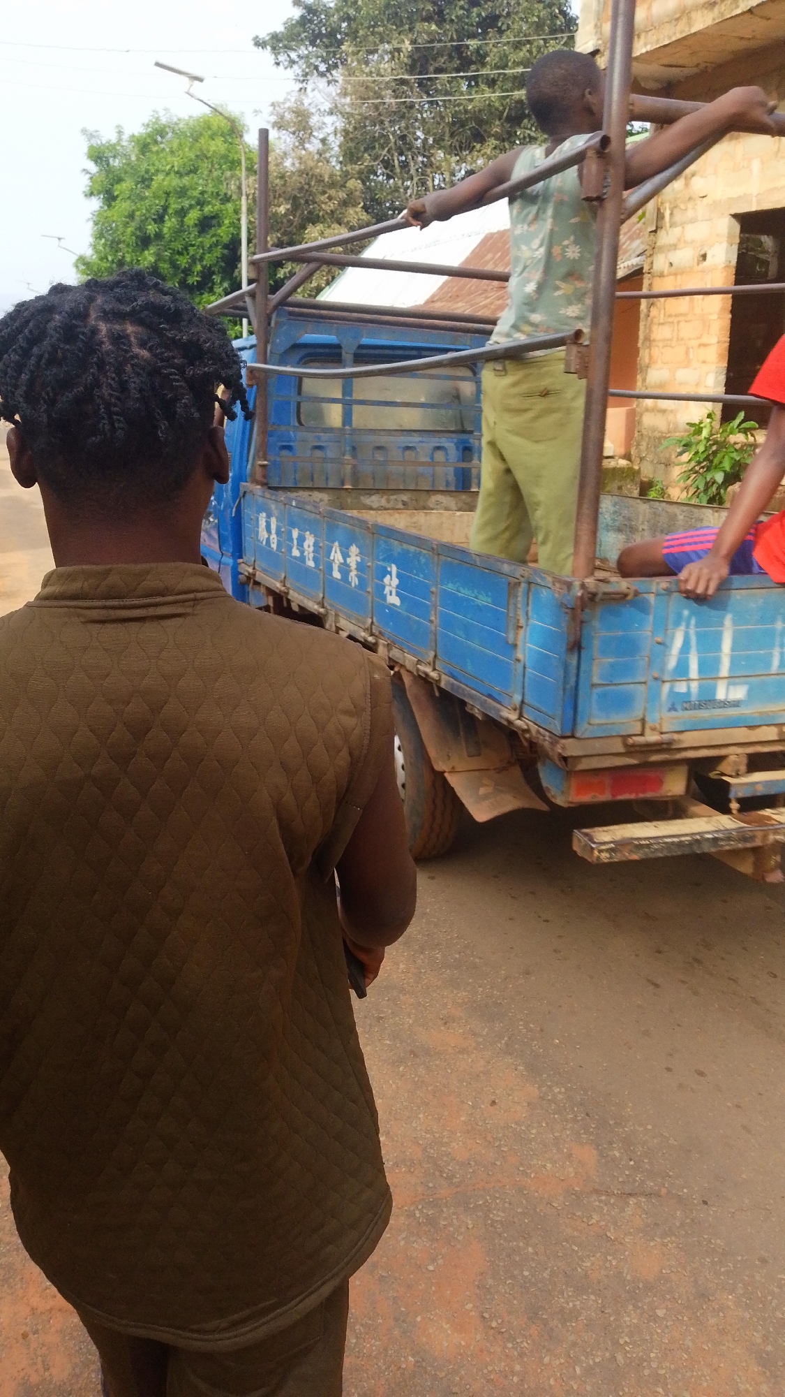 The image depicts a street scene in Enugwu-Ukwu, Nigeria. In the foreground, a person with braided hair wears a brown quilted vest and matching pants. The person's back is to the viewer. To the right, a blue flatbed truck with Chinese characters on its side is parked on the road. A young person stands in the truck bed, holding onto a metal frame. Another person sits at the back of the truck. The road is unpaved. A building is visible in the background. Trees are visible in the distance.