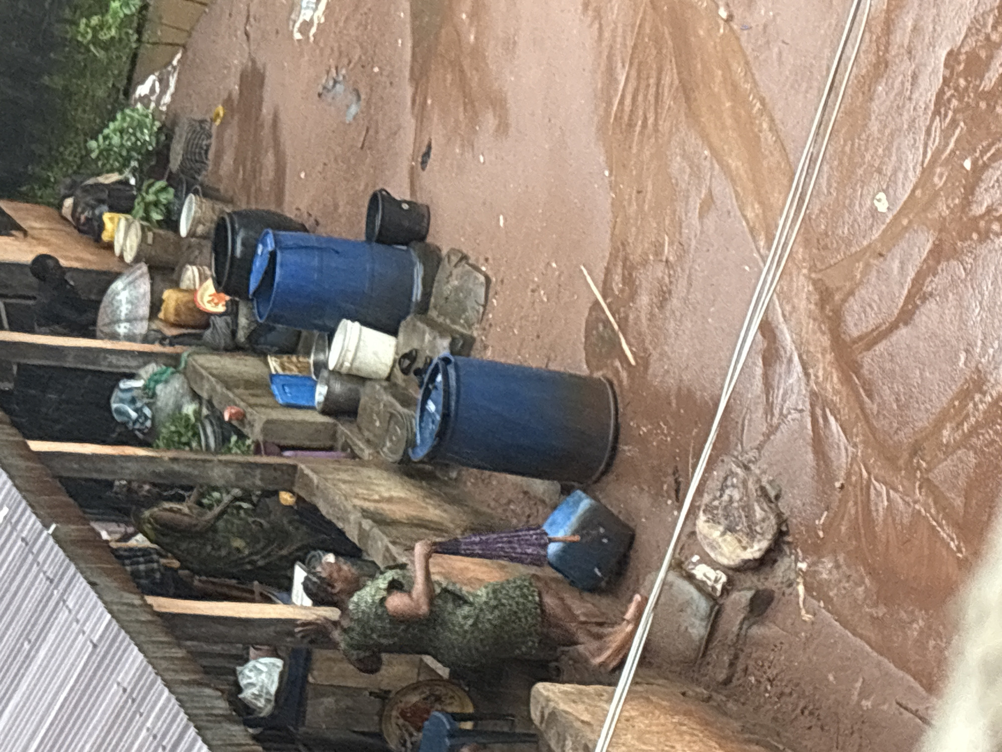 The image depicts a scene in Onitsha, Nigeria, during a downpour. In the foreground, a woman in a green, patterned dress is holding a closed, purple umbrella in her right hand and a blue plastic container in her left. Her bare feet are visible on the muddy ground. Behind her, a large blue plastic drum is situated next to a concrete wall. Further back, several other large containers and buckets are scattered around. A partially covered market stall with a corrugated metal roof is visible in the upper left of the frame, with some produce and other items stored within. The ground is a mixture of red mud and standing water, indicating significant rainfall.