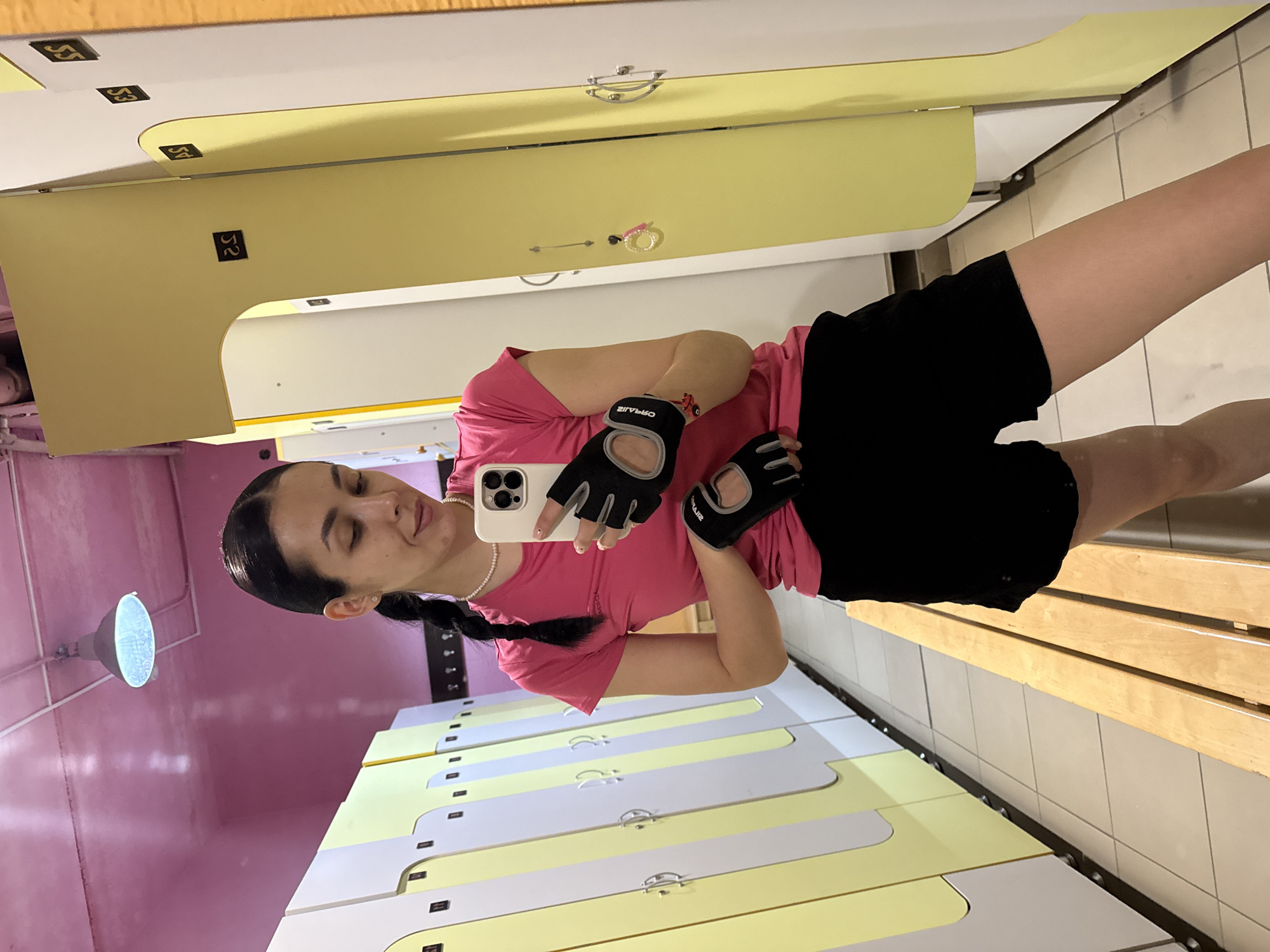 A woman is taking a selfie in what appears to be a locker room. She is wearing a pink t-shirt, black shorts, and workout gloves. She is holding a smartphone in her left hand. The background consists of rows of lockers, alternating between yellow and white. Above the lockers is a bench and a tiled wall. The ceiling and one wall are painted pink. A light fixture is visible on the ceiling. The location is potentially a gym or fitness center in Votkinsk, Russia, but that cannot be confirmed solely from the image.