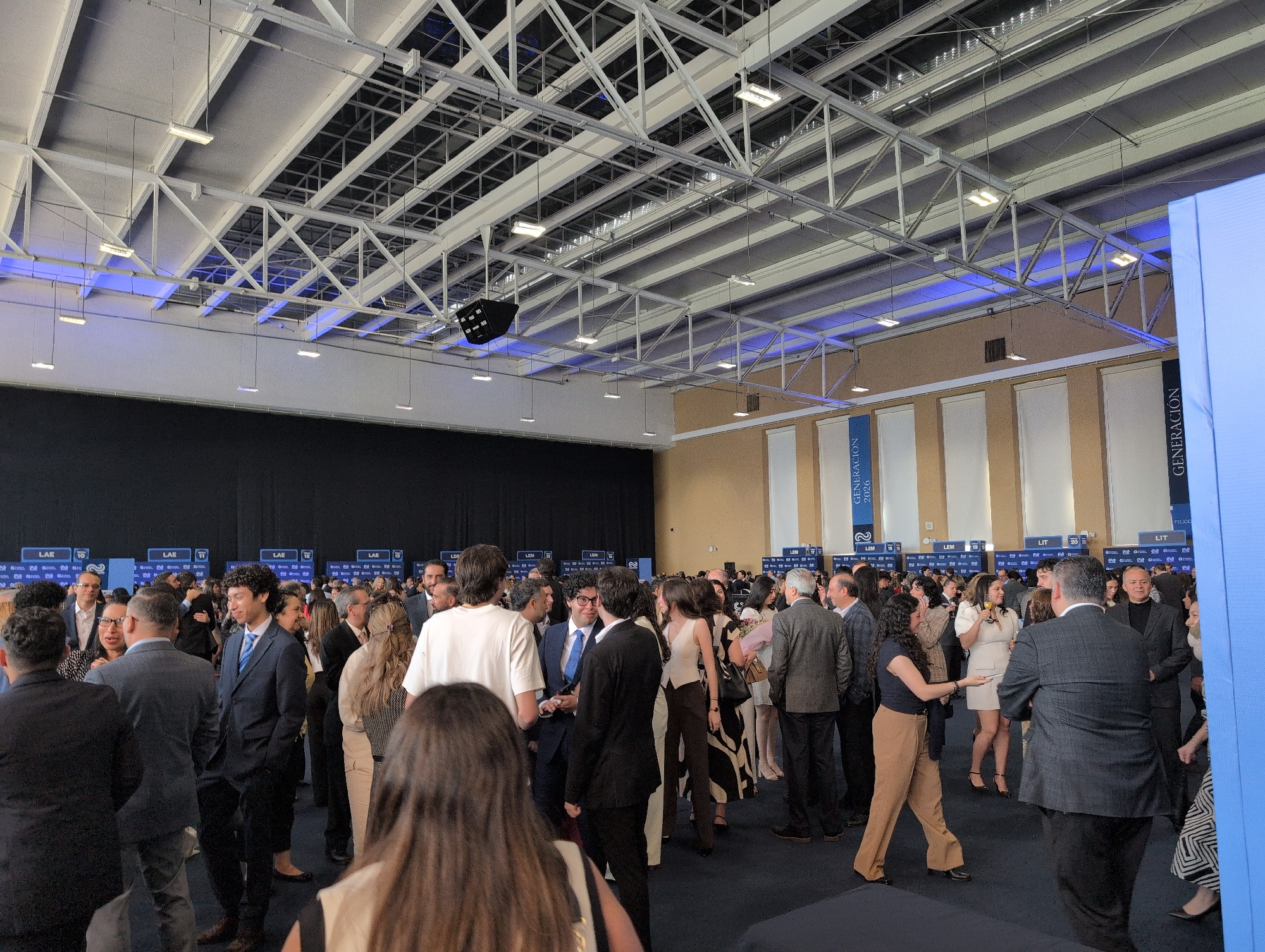The photograph captures an indoor event in Mexico City, Mexico, filled with a large crowd of people. Most are casually dressed in business attire such as suits, dresses, and blouses. They appear to be conversing in small groups. A black drape forms a backdrop on one side of the room, while several tall banners with the word "Generación" and the year "2026" are positioned along another wall. The venue is a large hall or conference center with a complex, interconnected ceiling structure featuring recessed lighting, giving the space a modern architectural aesthetic. The room appears to be well-lit and is set up for what seems to be a professional gathering or convention.