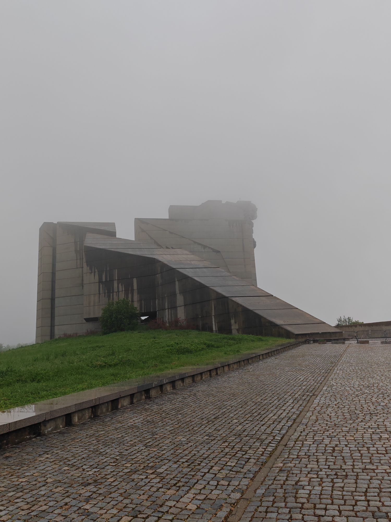 The image shows the Monument to 1300 Years of Bulgaria, located in Shumen, Bulgaria. The monument is constructed of concrete and depicts stylized figures. A sloped walkway leads up to the monument, and the ground is covered with stone blocks. The sky is overcast with heavy fog.
