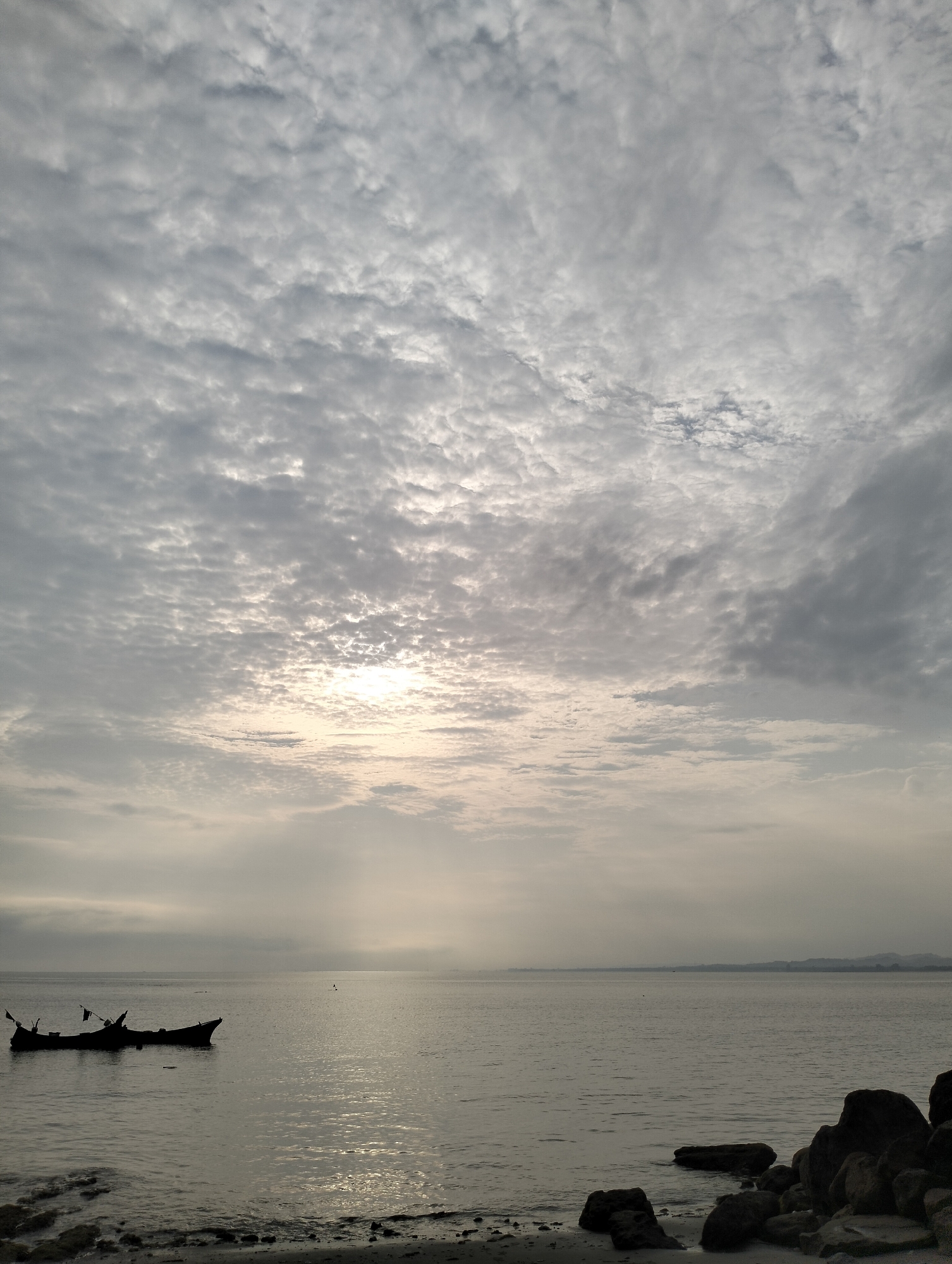 The image depicts a seascape under a cloudy sky, likely at dawn or dusk. A dark silhouette of a small, possibly damaged, boat is visible on the water to the left. The shoreline in the foreground features rocks and sand. In the distance, faintly visible land suggests a coastline. The sun is behind the clouds, creating a diffused light effect. The scene is somber and still. No people are visible. It is likely in Lhokmanee, Indonesia.