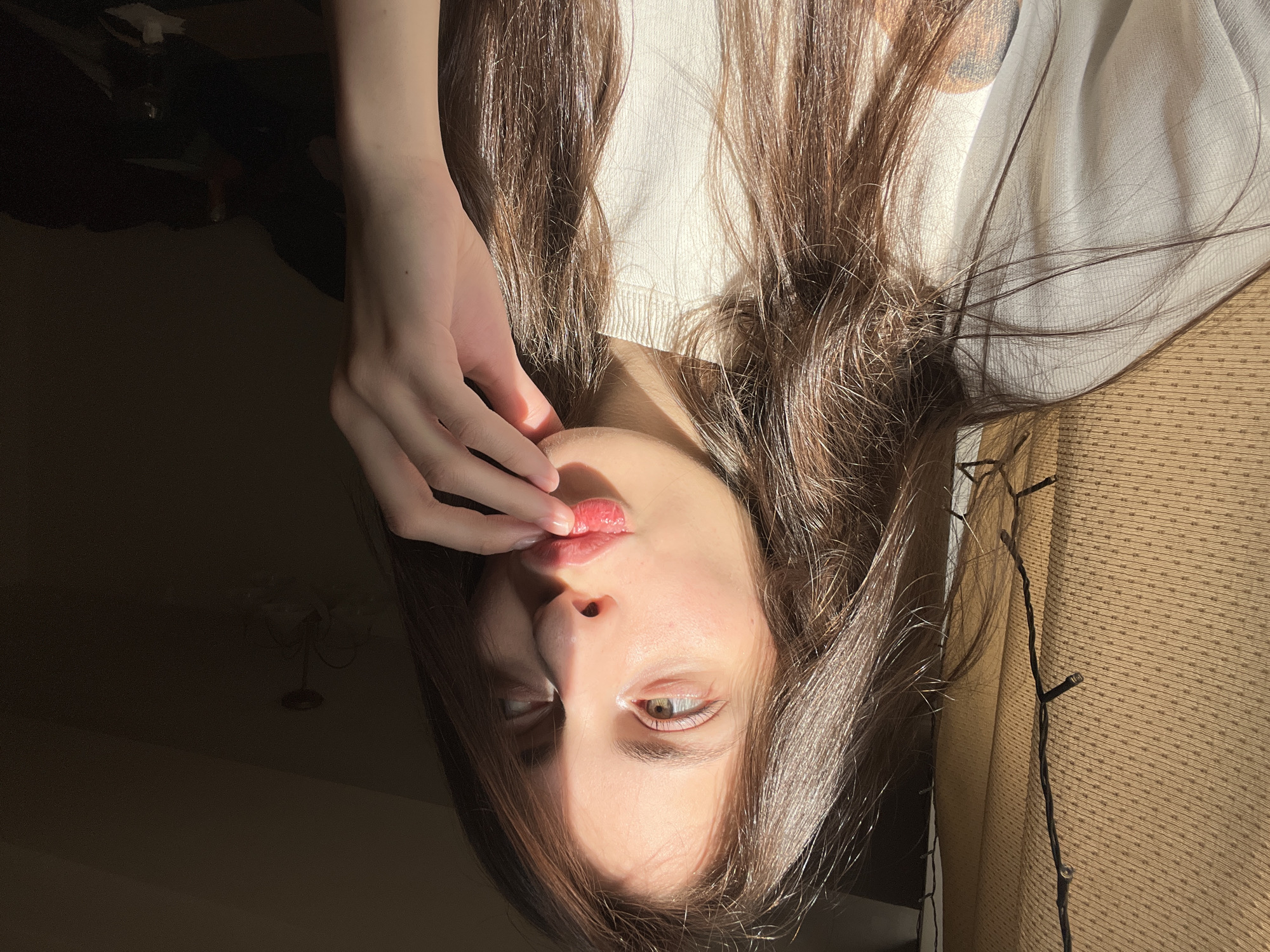 A young woman with long brown hair is seen in a head-and-shoulders shot, inverted. She is wearing a light-colored top. Her hand is near her lips. The background includes a dark space, possibly a room, and a lighter area with textured material and what appear to be string lights. There is no evidence to suggest the location is Krasnoyarsk, Russia.