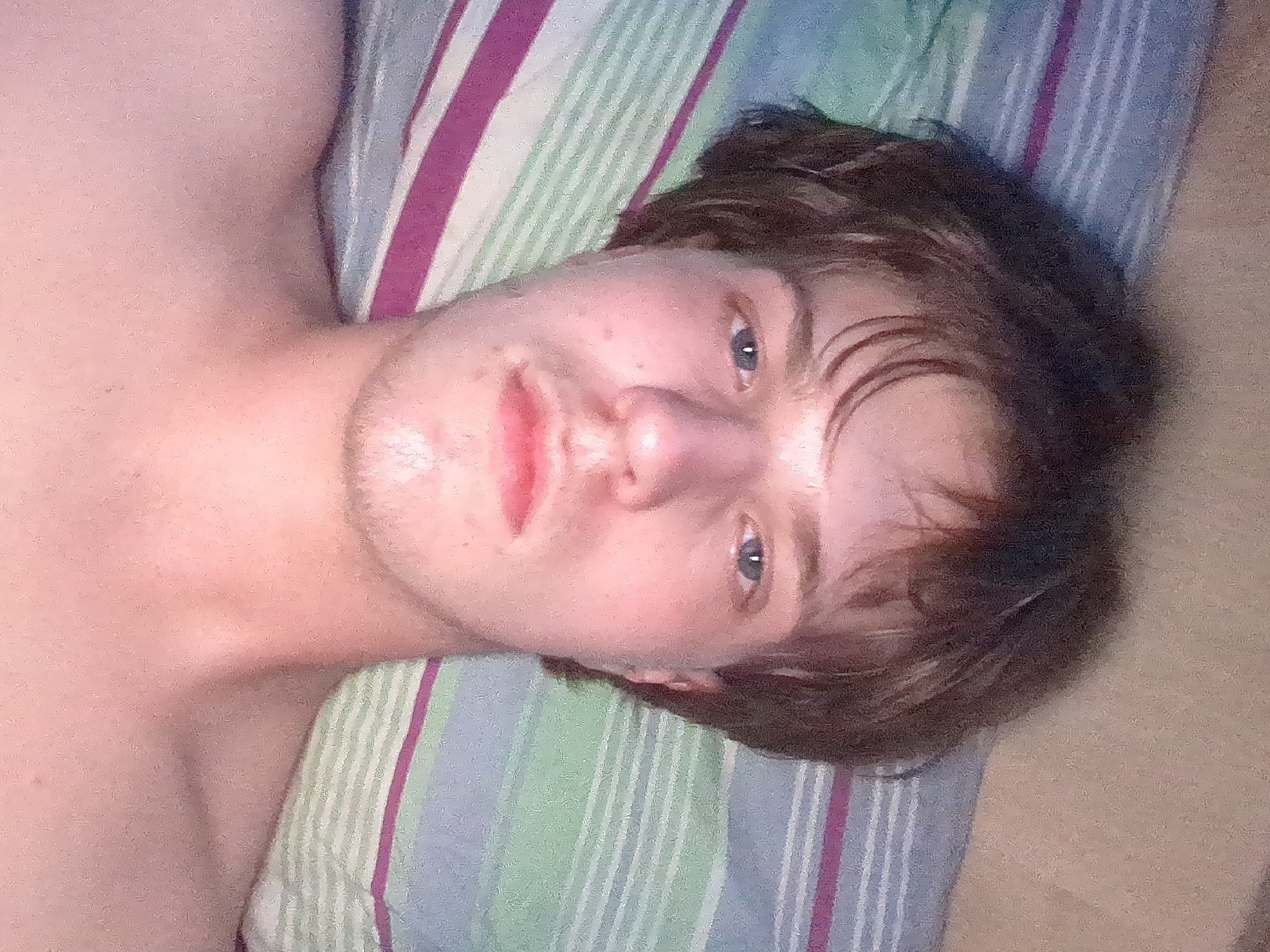 The image is a close-up, selfie-style photograph of a young person lying down on a striped pillow. The person has light skin, light blue eyes, and medium-length, light brown hair. They are looking directly at the camera. The background consists of the pillow, which has horizontal stripes in shades of blue, green, and purple. The lighting appears to be artificial, possibly from overhead. The image quality is slightly grainy. There is no indication of the location being Votkinsk, Russia.