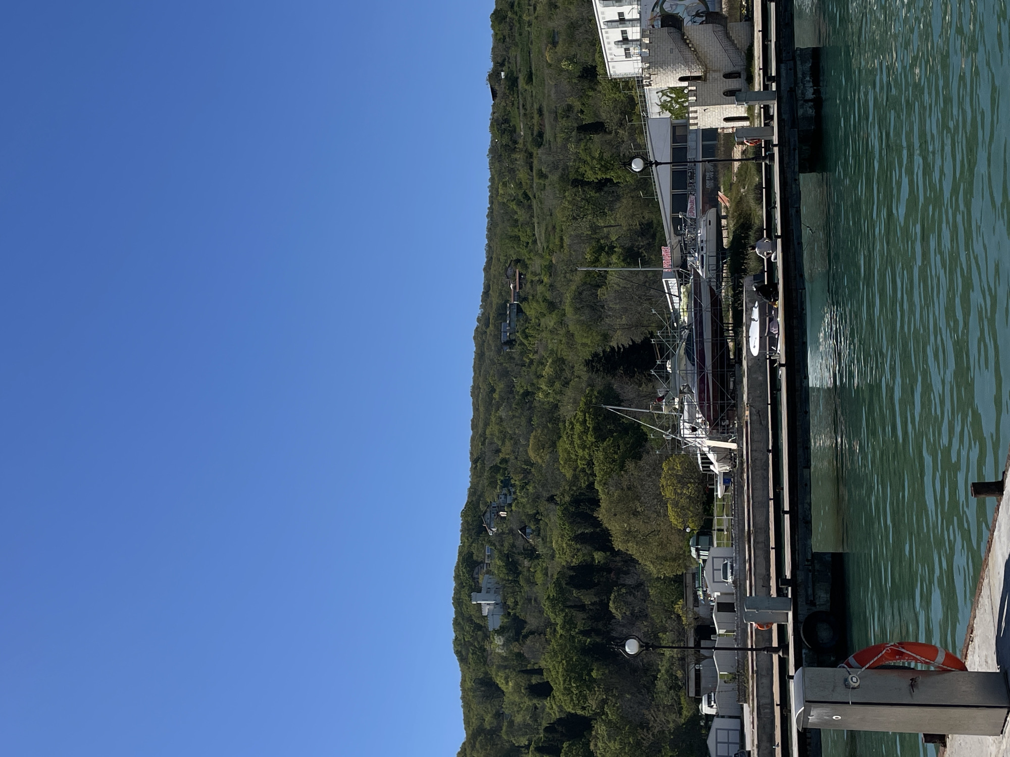 The image shows a waterfront scene in Varna, Bulgaria. A blue sky fills the left side of the image. A green, tree-covered hillside slopes down towards the water's edge on the right. At the edge of the water is a dock or pier with boats moored alongside. There are buildings among the trees on the hillside and more buildings near the dock. A life preserver is visible on the edge of the dock. There are no people obviously visible in the image. The water appears calm. The scene is likely a marina or harbor area.