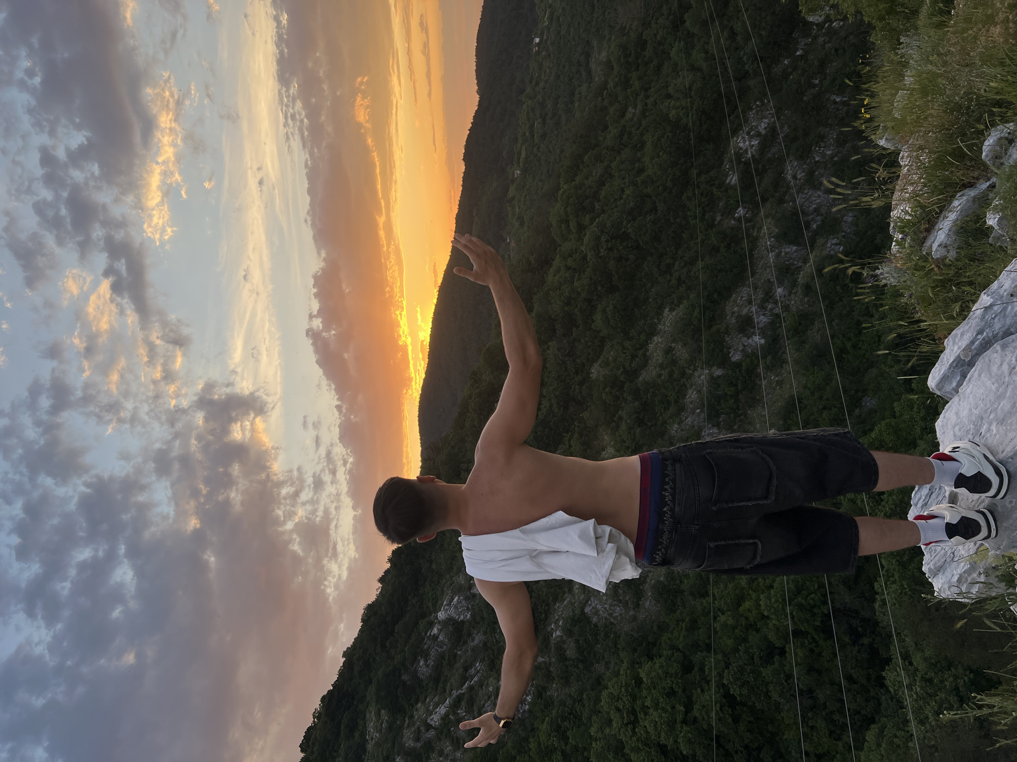 A man stands on a rocky outcrop overlooking a green, mountainous landscape against a backdrop of a colorful sunset. The man is facing away from the camera, shirtless with a white t-shirt draped over his back and shoulders. He is wearing dark shorts and white and black sneakers with red accents on the socks. His arms are outstretched, one raised towards the sky and the other lowered to his side. The sky exhibits a gradient of colors from shades of orange near the horizon to blues and grays higher up, with clouds scattered throughout. The scene is possibly at or near a viewpoint within the Asenovgrad area of Bulgaria, given the terrain, vegetation, and sunset, and it is rotated clockwise by ninety degrees. Overhead power lines may be seen in the background.