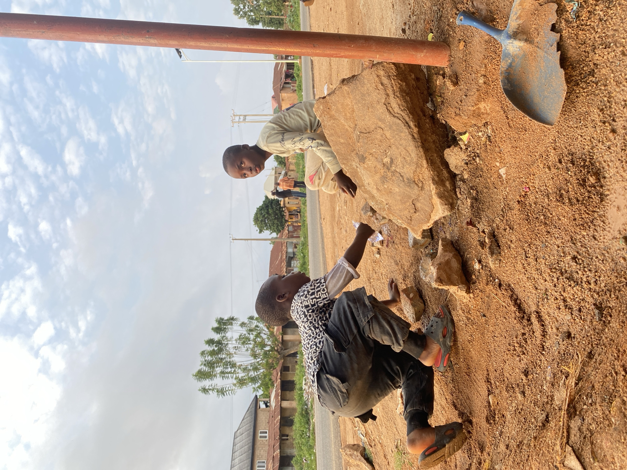 Two young boys are positioned near a large rock formation and a pole. One boy, wearing a light-colored long-sleeved shirt, stands and looks down while leaning against the rock. The other boy, wearing a patterned shirt and dark pants, is crouched on the ground, interacting with something near the rock. A shovel is present near the rock. The ground consists of a reddish-brown soil. Buildings are visible in the background. The image was taken in Vom, Nigeria.