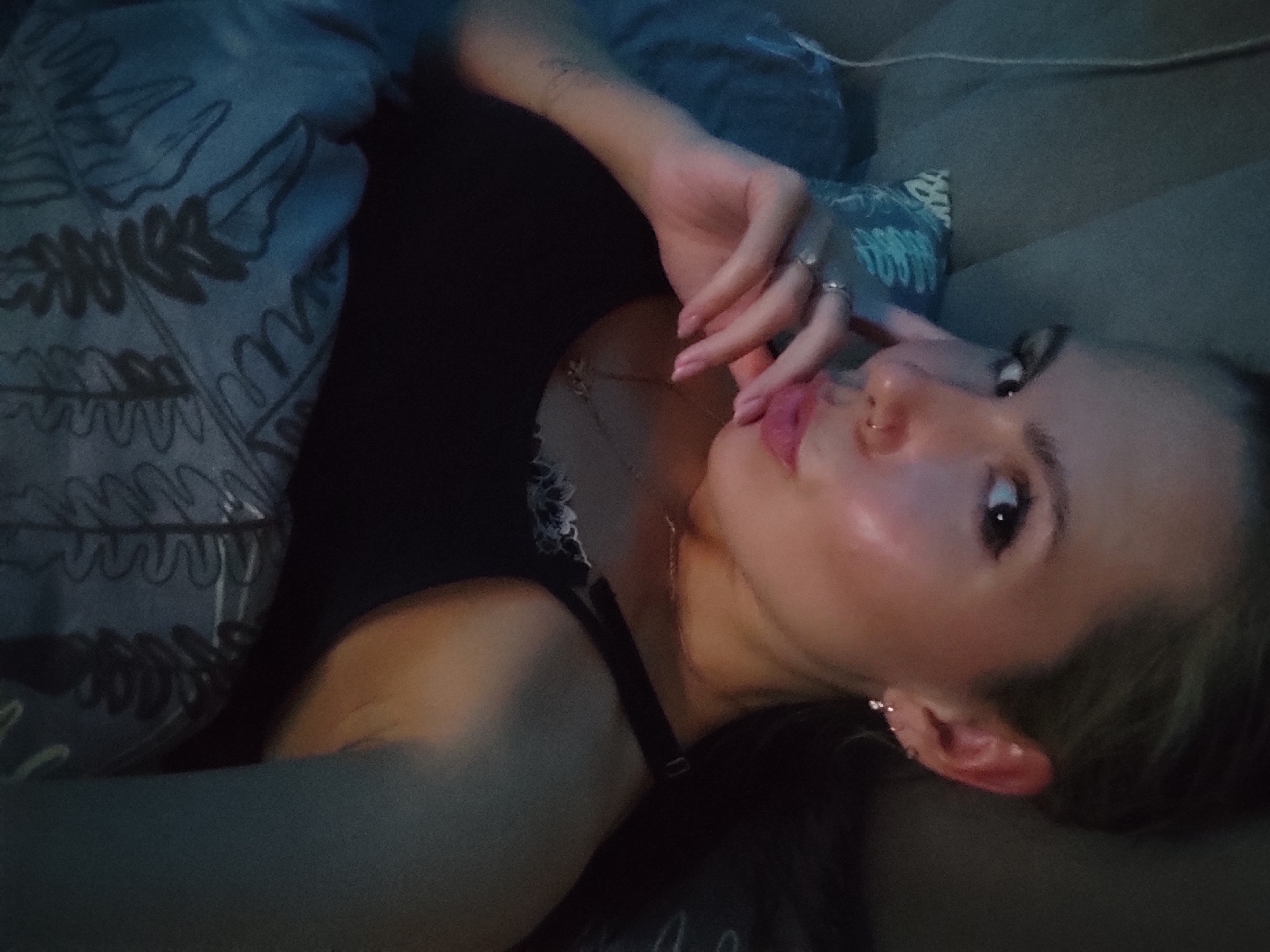 A young woman lies on a neutral-colored couch. She has fair skin, pink lips, and brown eyes. Her left hand is raised near her face. She wears a silver necklace and earrings. She is wearing a black top with a white floral pattern on the edges. A patterned blue fabric is visible to the left of her. The location is the city of Chaikovsky, Russia.