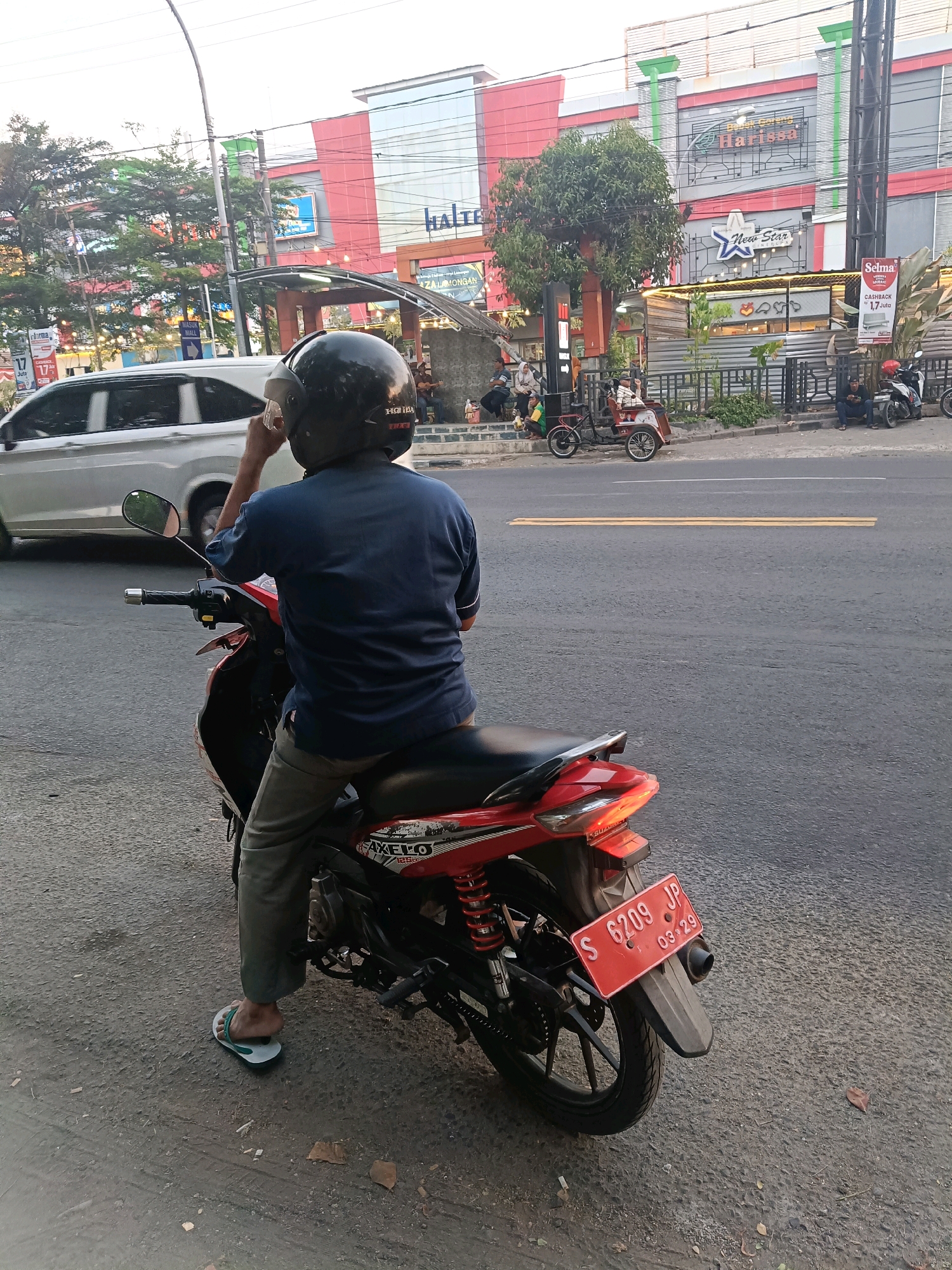 A person wearing a dark blue shirt and grey pants is sitting on a red and black motorcycle, looking towards a building with the sign "Halte" and "Harissa" in Lamongan, Indonesia. The person is wearing a black helmet and white flip-flops. The motorcycle has a red license plate with the number "S 6209 JP" and "03 29" on it. In the background, there is a street with a white car, several other motorcycles, a few people sitting on steps, and a trike. The building appears to be a shopping center or mall with multiple shops. The scene is captured during daylight with clear weather.